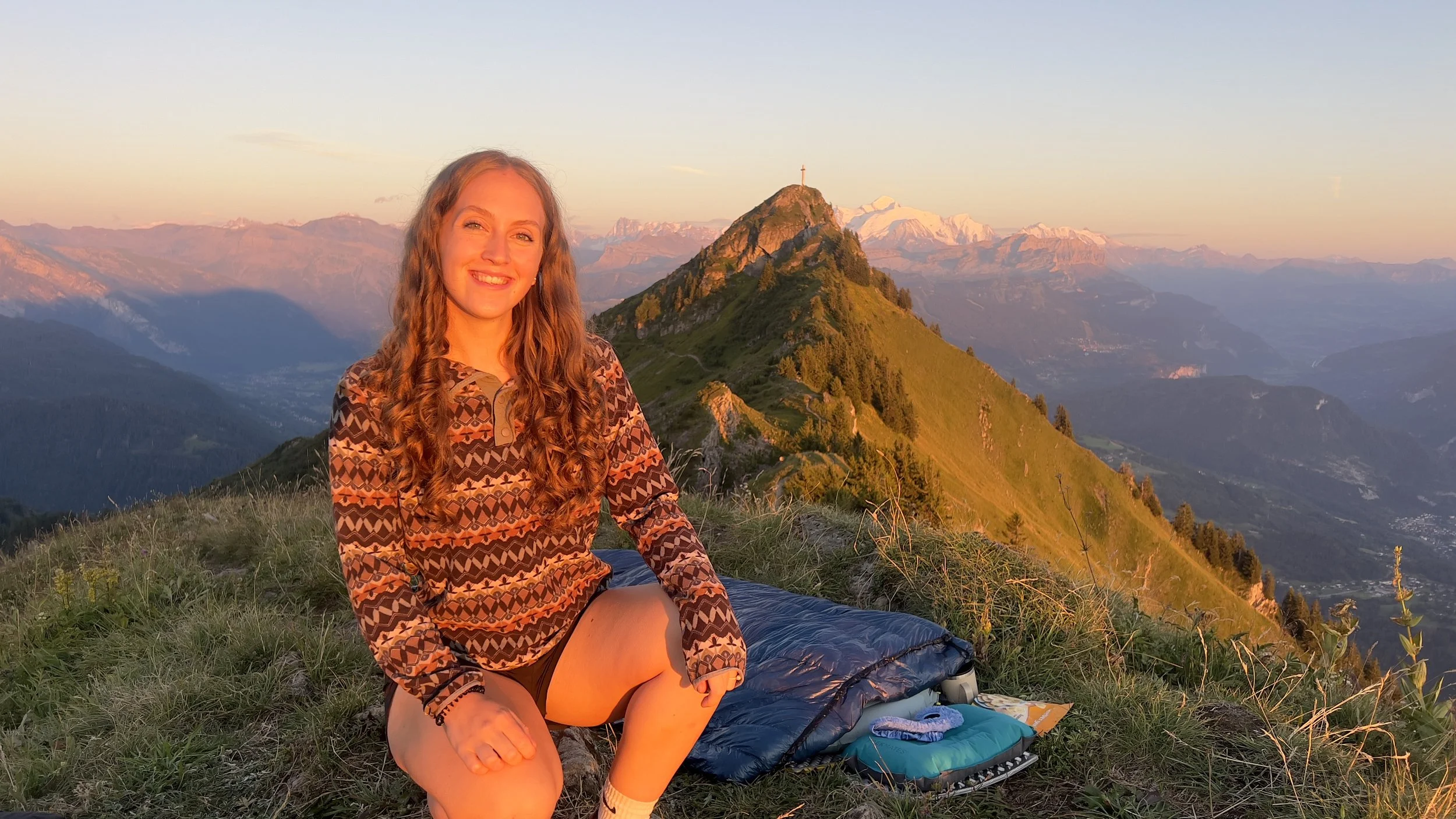 A young woman with long curly hair smiling, sitting on grass with a mountain range in the background during sunset.