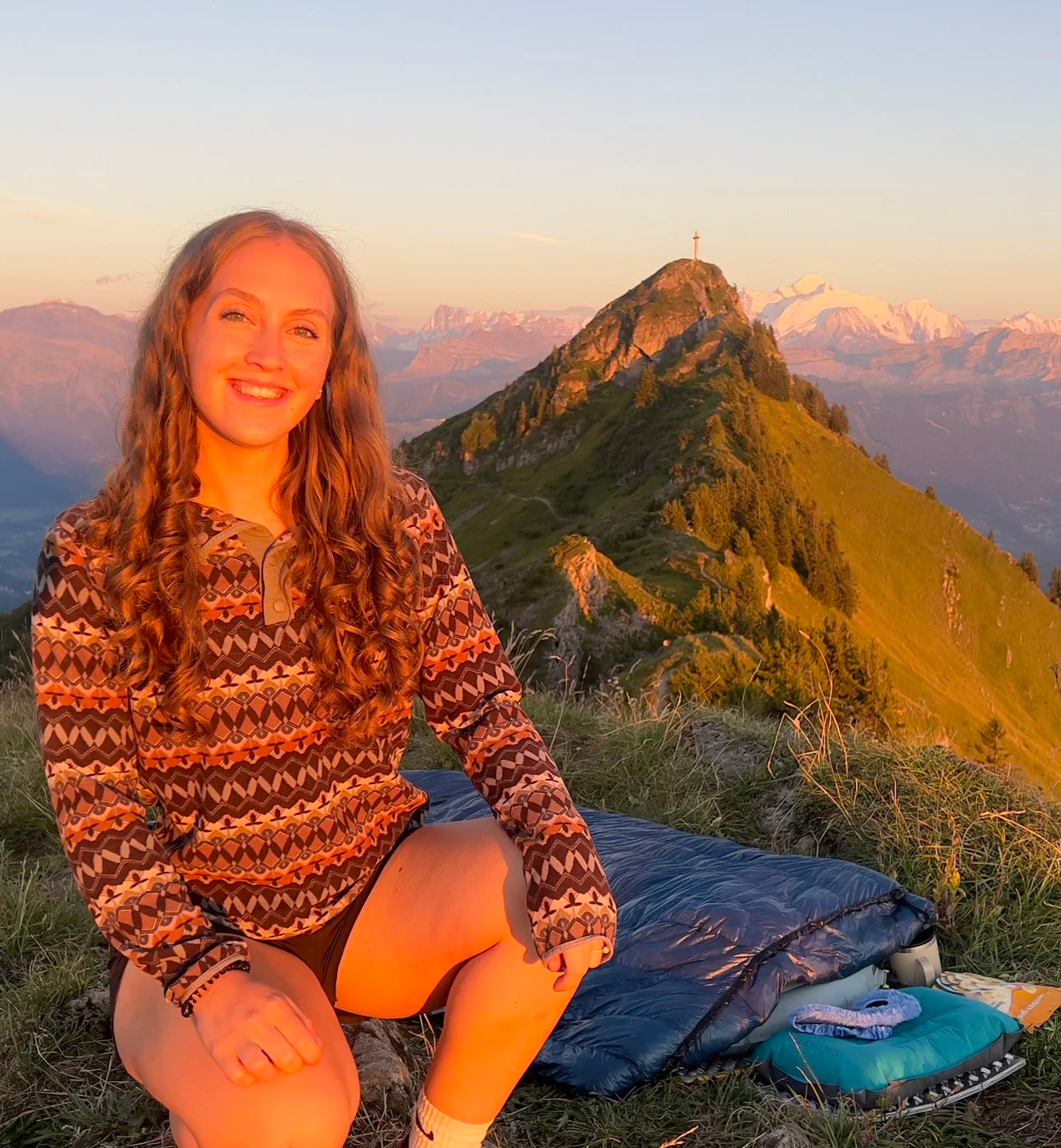 A young woman with long, curly hair smiling at sunset outdoors, sitting on a grassy hillside with mountains in the background, near camping gear.