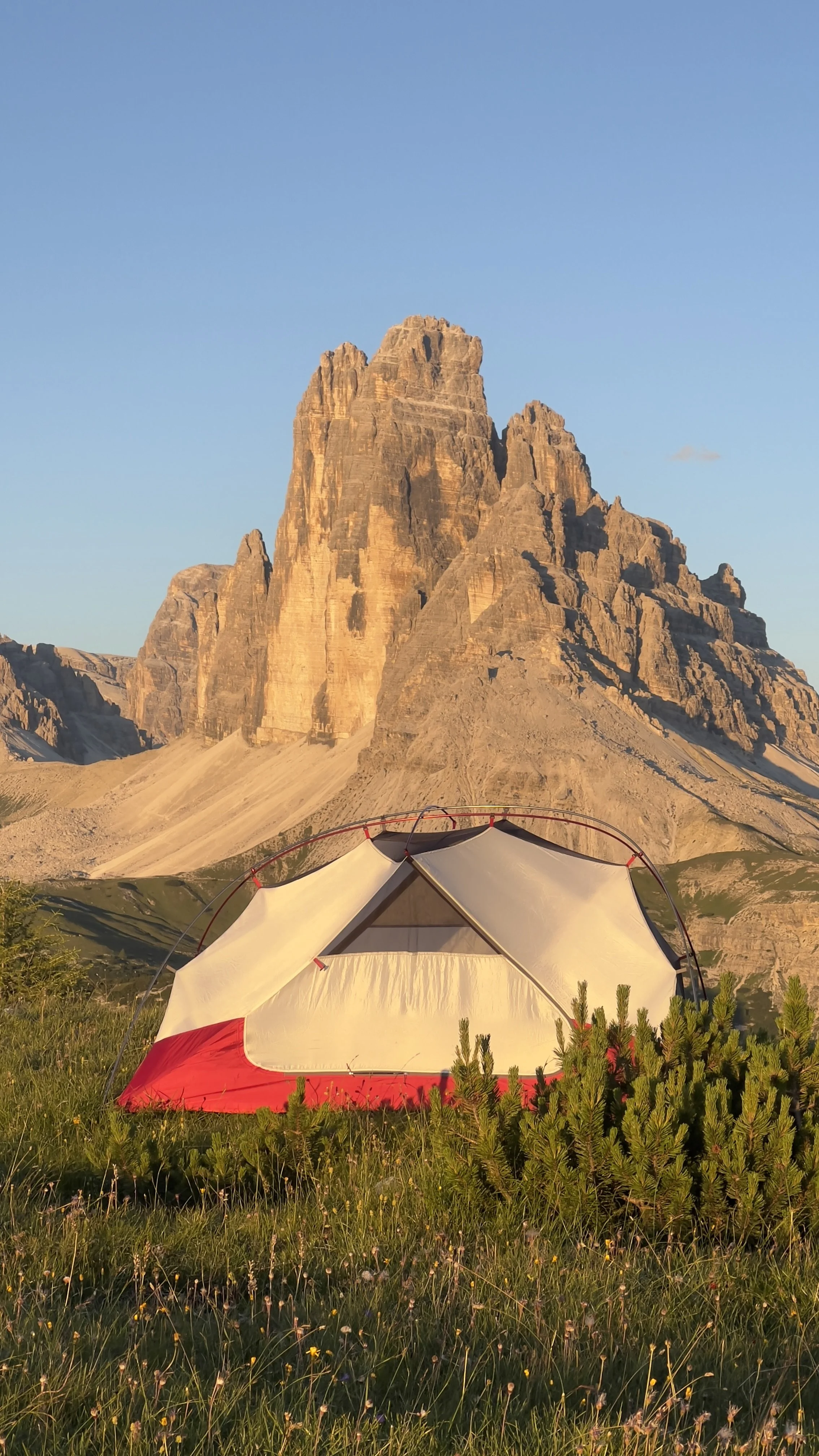 A camping tent set up in a grassy field with green bushes in front of a large mountain, likely the Tre Cime di Lavaredo, in the Dolomites during sunset.