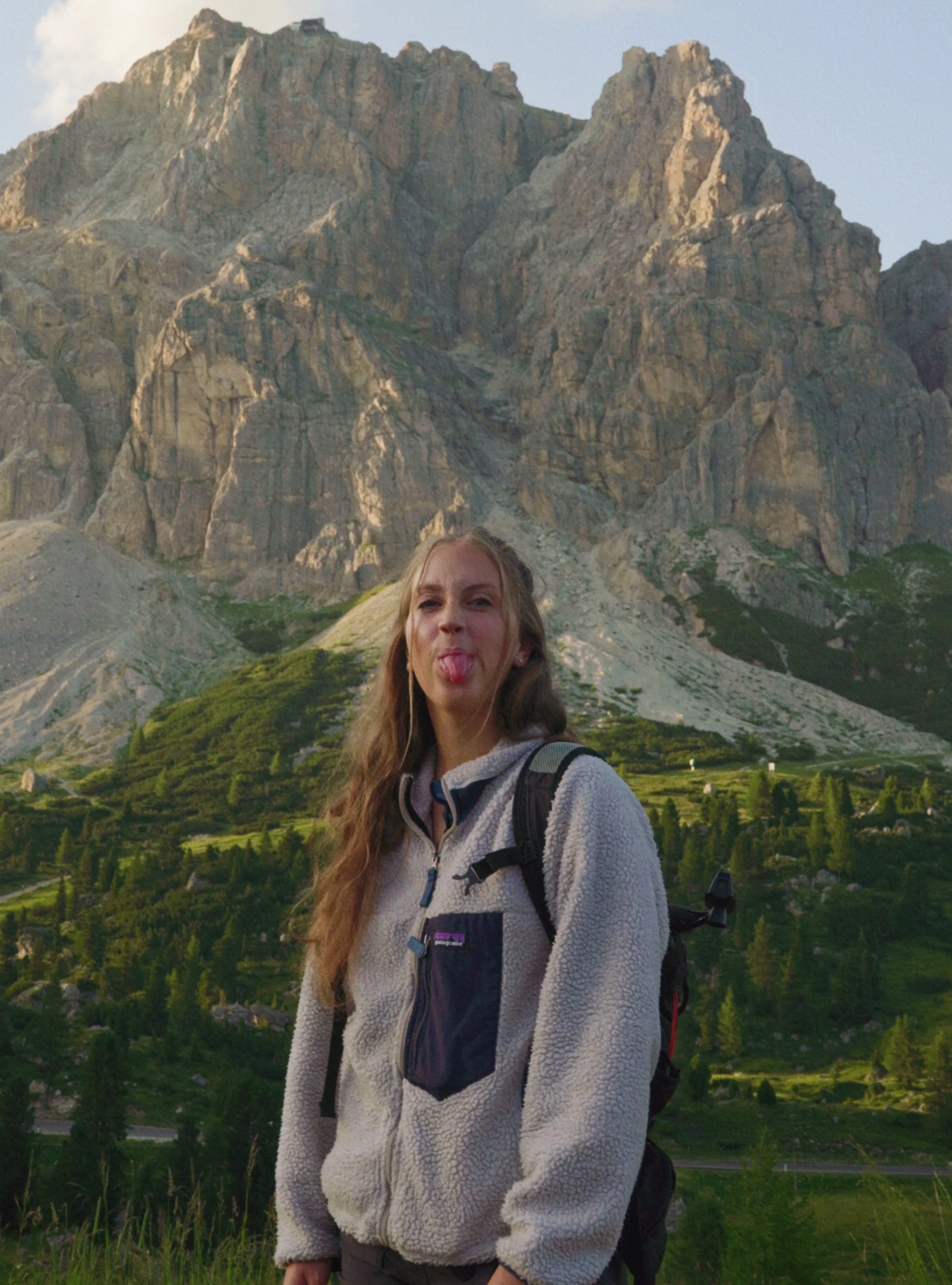A young woman with long brown hair sticking out her tongue, standing outdoors with a mountain range in the background. She is wearing a light-colored fleece jacket and a backpack.