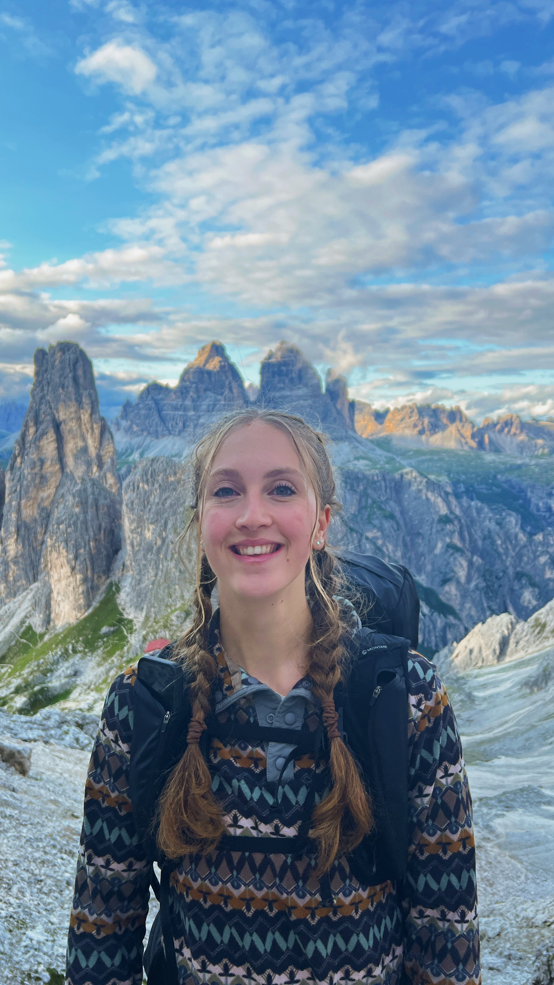A young woman with braided hair, wearing a patterned jacket and carrying a backpack, smiling outdoors with mountains and a partly cloudy sky in the background.