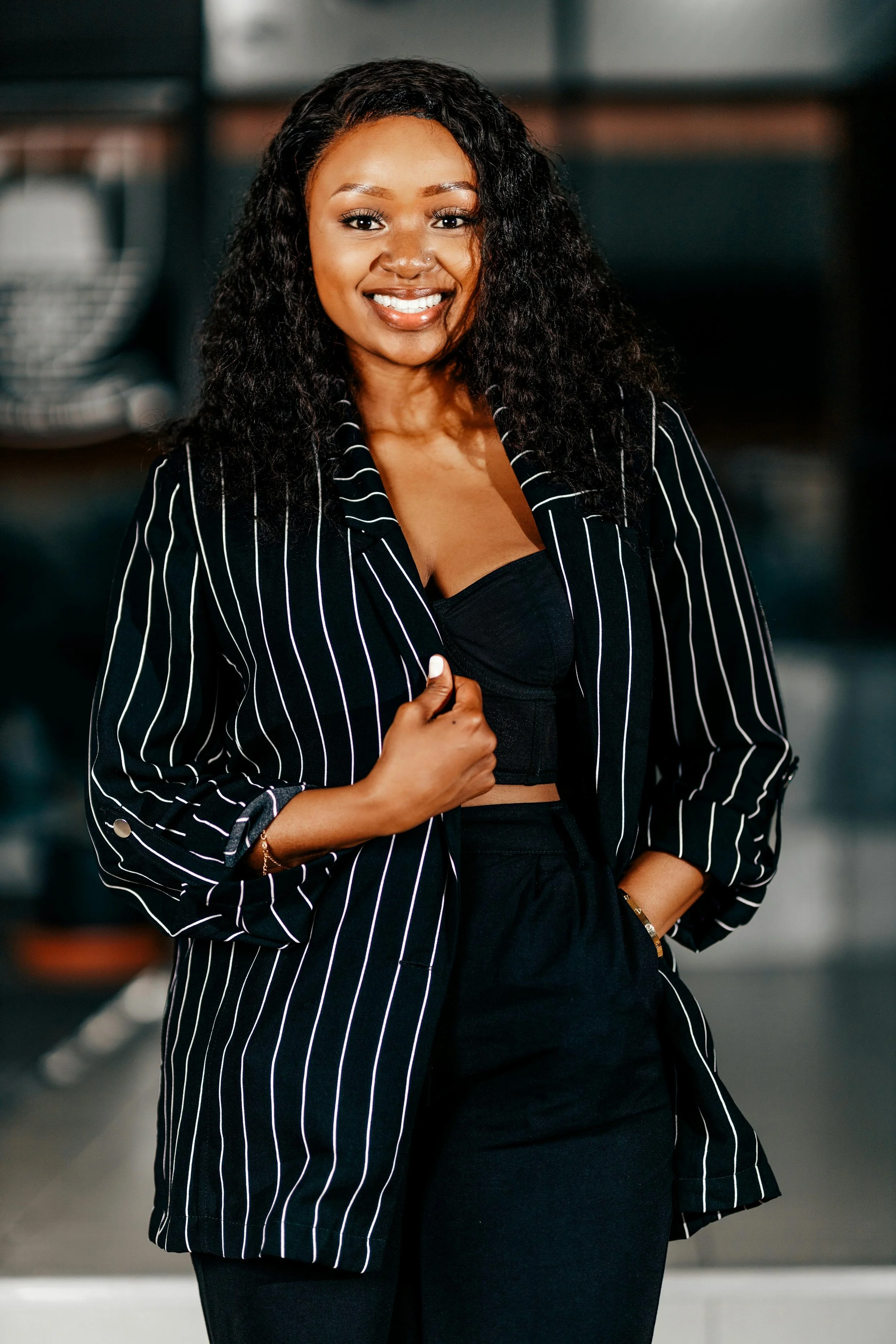 A confident woman with curly black hair, wearing a black top and a black and white pinstripe blazer, smiling and standing with her hand on her chest. The background appears to be an urban setting.