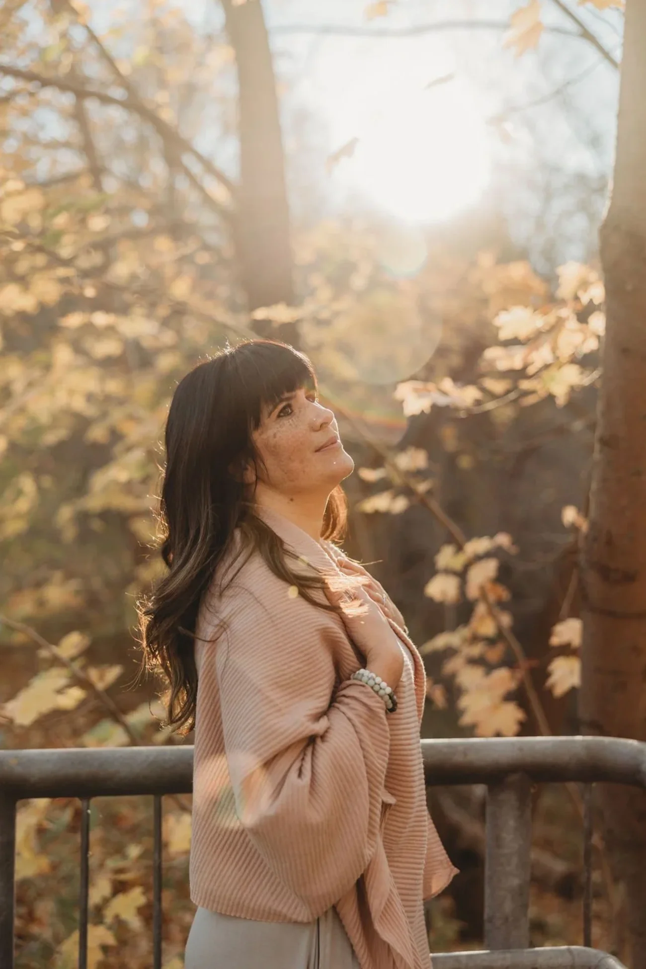 Laura of Healing With Love in Hamilton wearing a light pink jacket and bracelets, standing outdoors in a wooded area during sunset, with her hand on her chest and gaze upwards.
