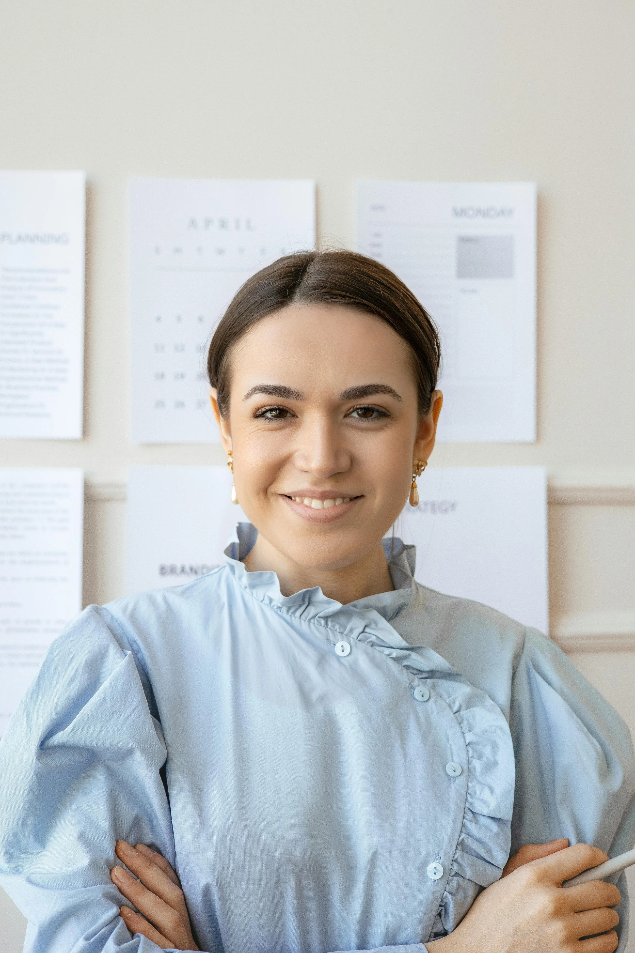 Smiling woman in medical gown with arms crossed standing in front of a wall with printed calendar pages.