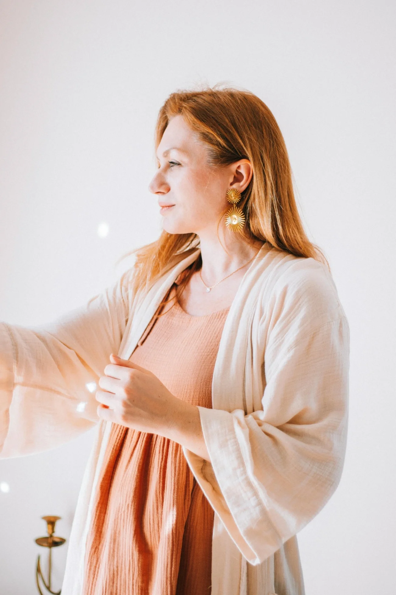A woman with reddish hair wearing earrings and a layered peach-colored dress under a cream-colored cardigan, holding a string of lights against a white background.