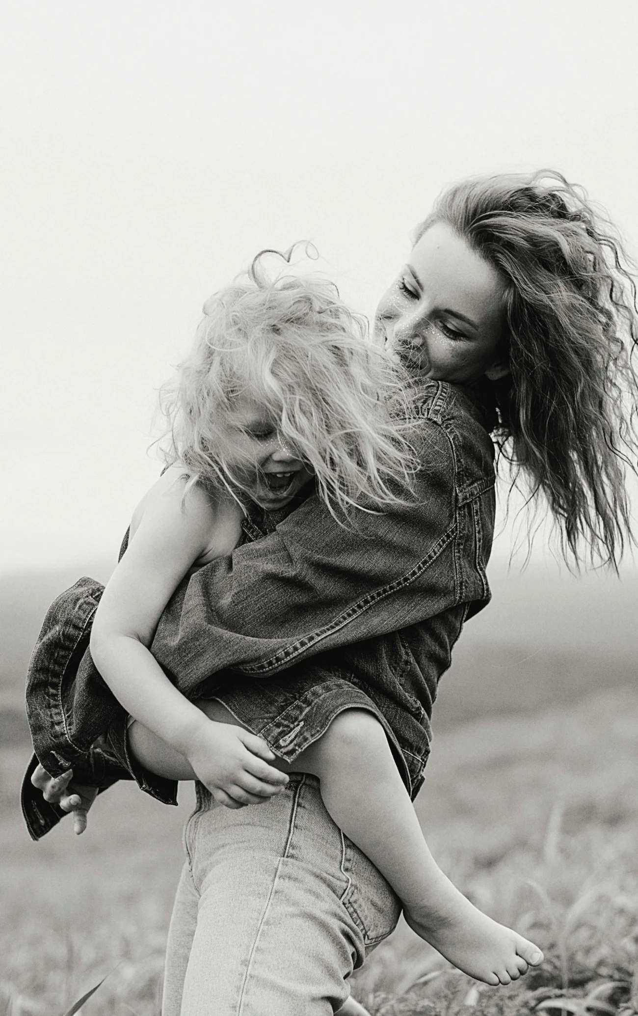 A woman with curly hair hugging and smiling at a child with curly hair, both appear joyful, outdoors in a grassy field, black and white photo.