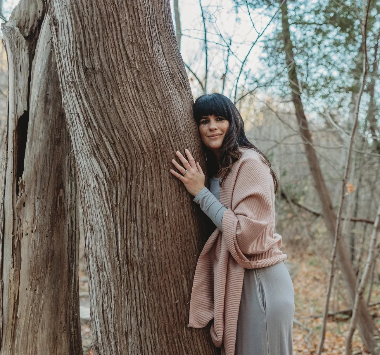 Laura of Healing With Love in Hamilton wearing a light pink sweater and beige pants, hugging a large tree trunk in a forest setting during fall.