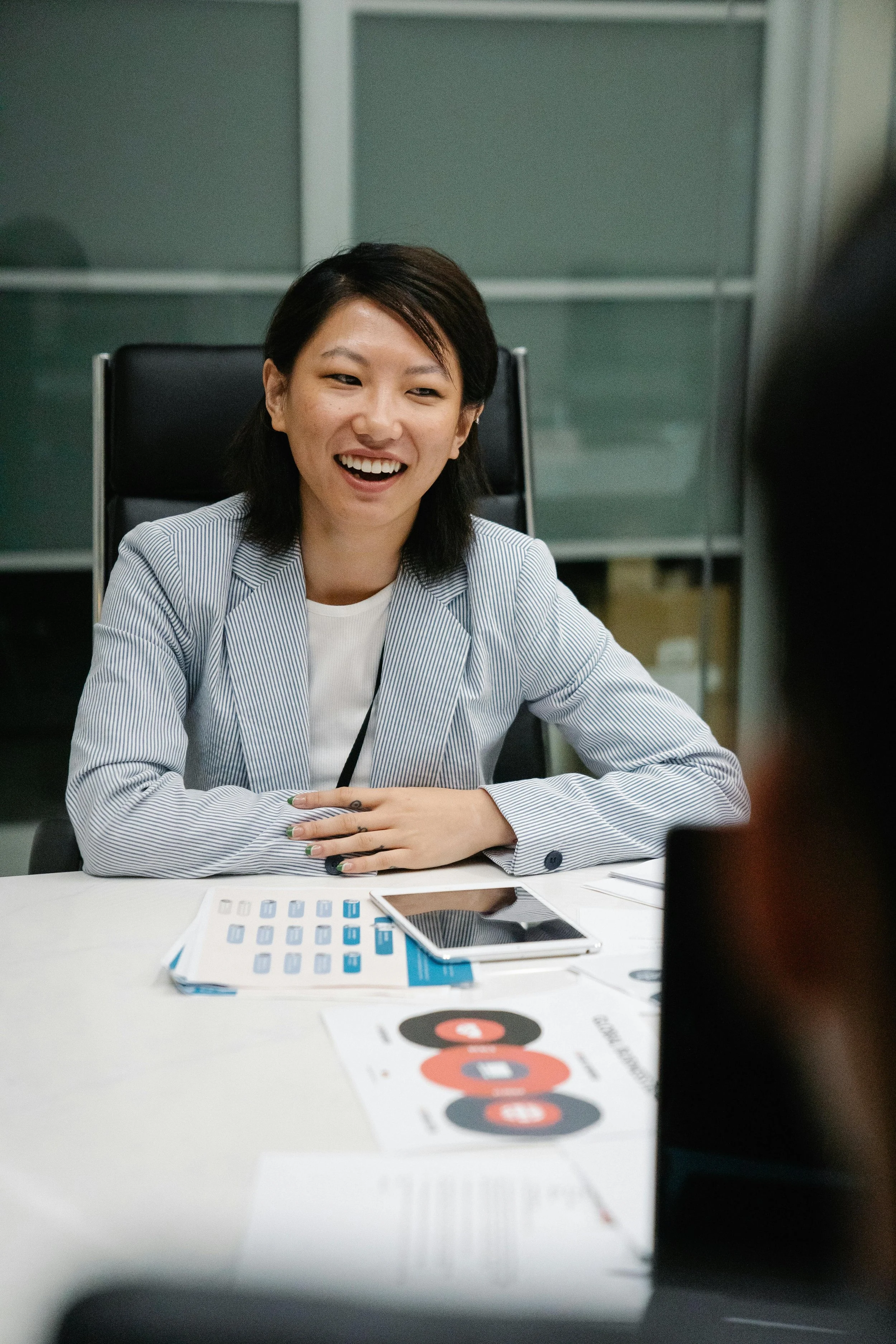A woman with short dark hair is smiling and talking during a meeting. She is sitting at a white table with a tablet, printed charts, and a smartphone in front of her. The background has a modern office setting with frosted glass panels.