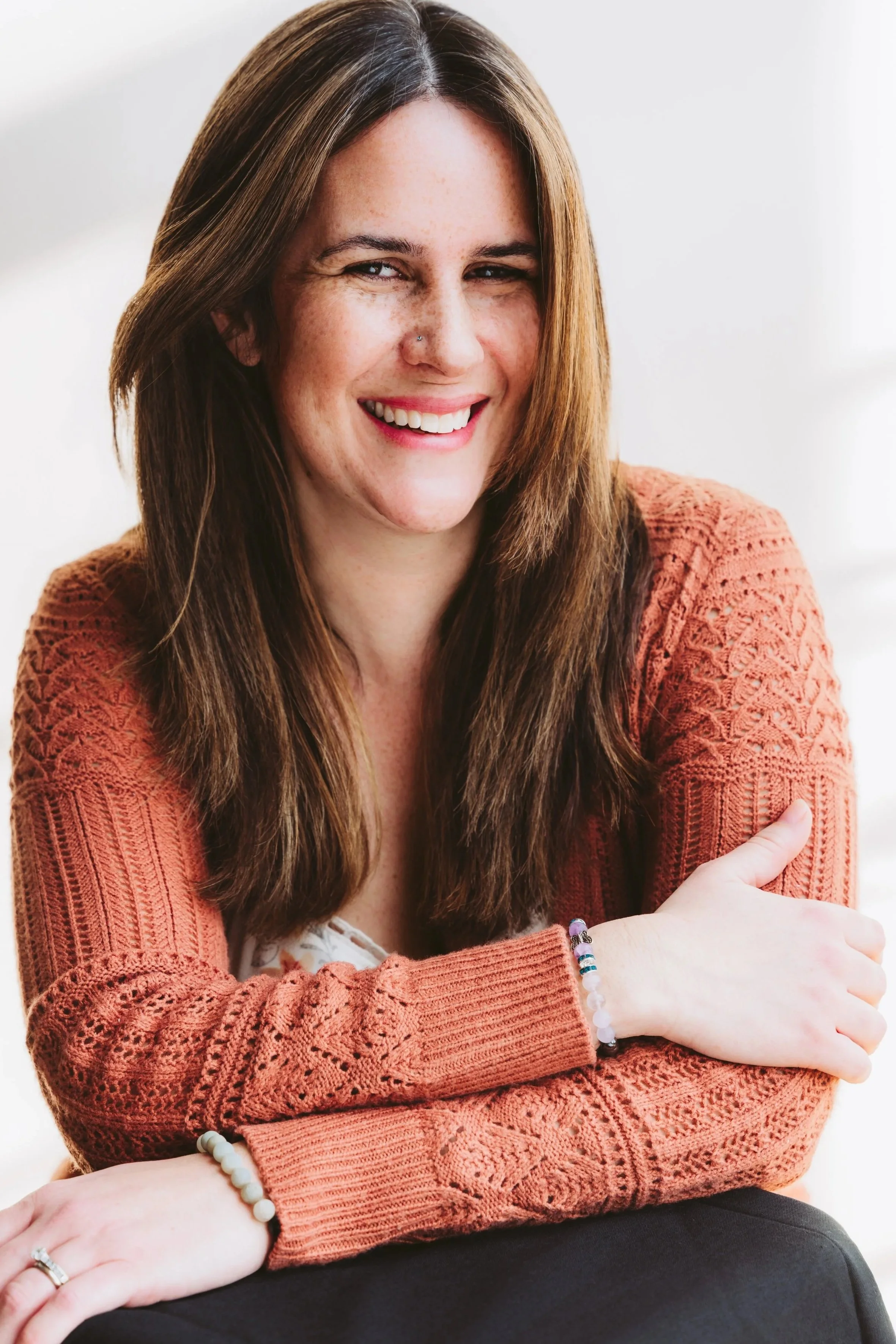 A woman with long brown hair, smiling, wearing a rust-colored lacy sweater, bracelets, and rings, sitting with crossed arms against a light background.