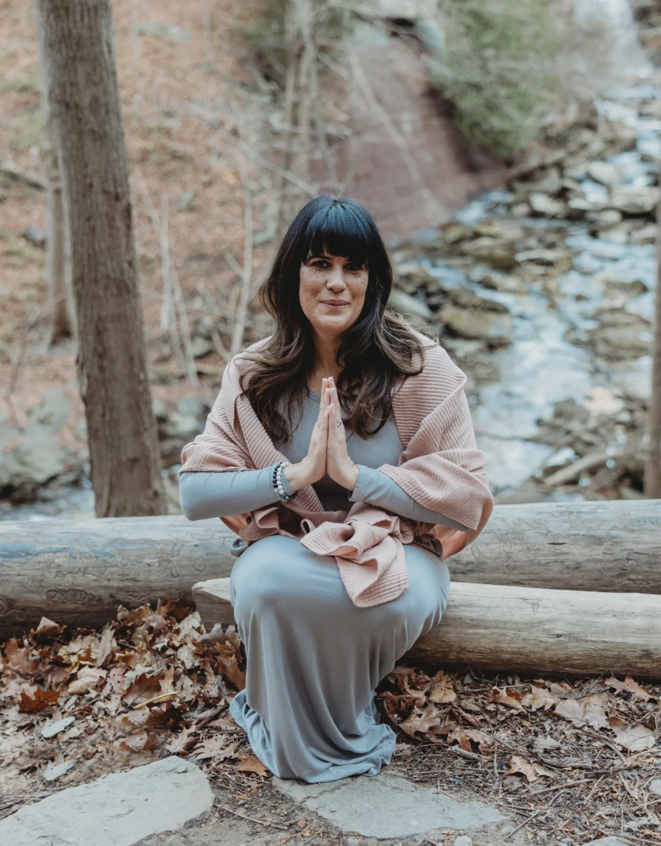 Laura of Healing With Love in Hamilton in a seated pose making a prayer gesture with hands pressed together, sitting on a fallen log in a forested area near a stream.
