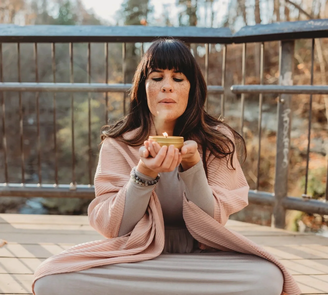 Laura of Healing With Love in Hamilton sitting outdoors on a wooden platform, holding a small round container with a lit candle or incense, with her eyes closed, surrounded by trees in the background.