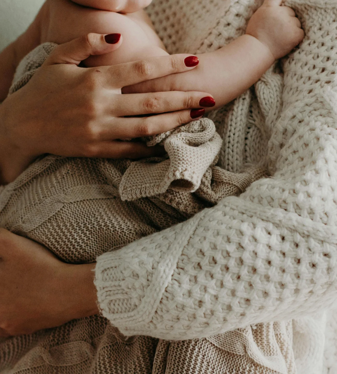 Woman's hand holding a newborn baby's arm, with the baby wrapped in knitted and woven blankets.