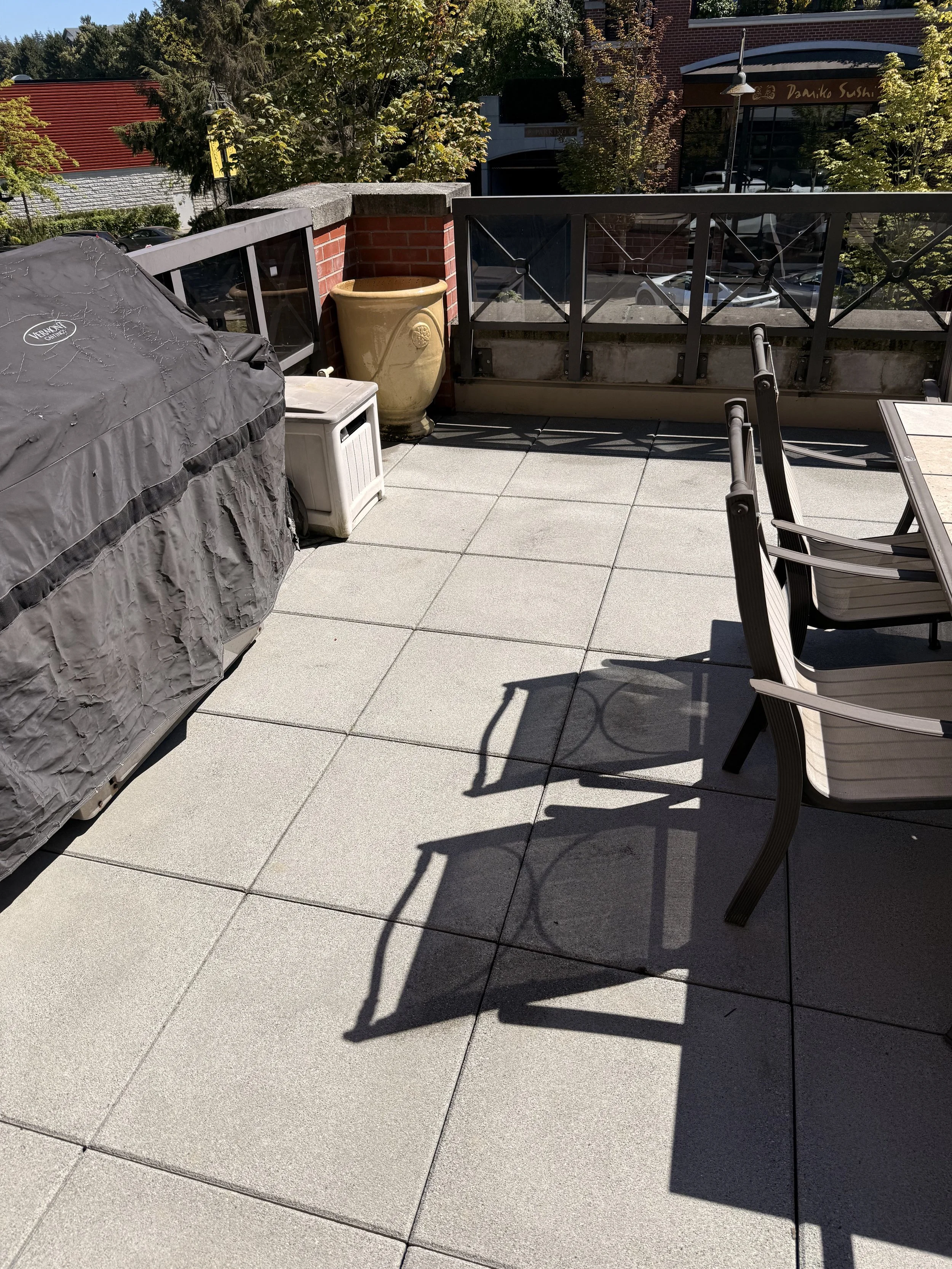 View of a balcony or rooftop patio with outdoor furniture, including chairs and a covered grill, a large yellow ceramic planter, a small white side table, and a decorative brick wall with an aluminum railing. In the background, trees, buildings, parked cars, and a sign for a sushi restaurant are visible.
