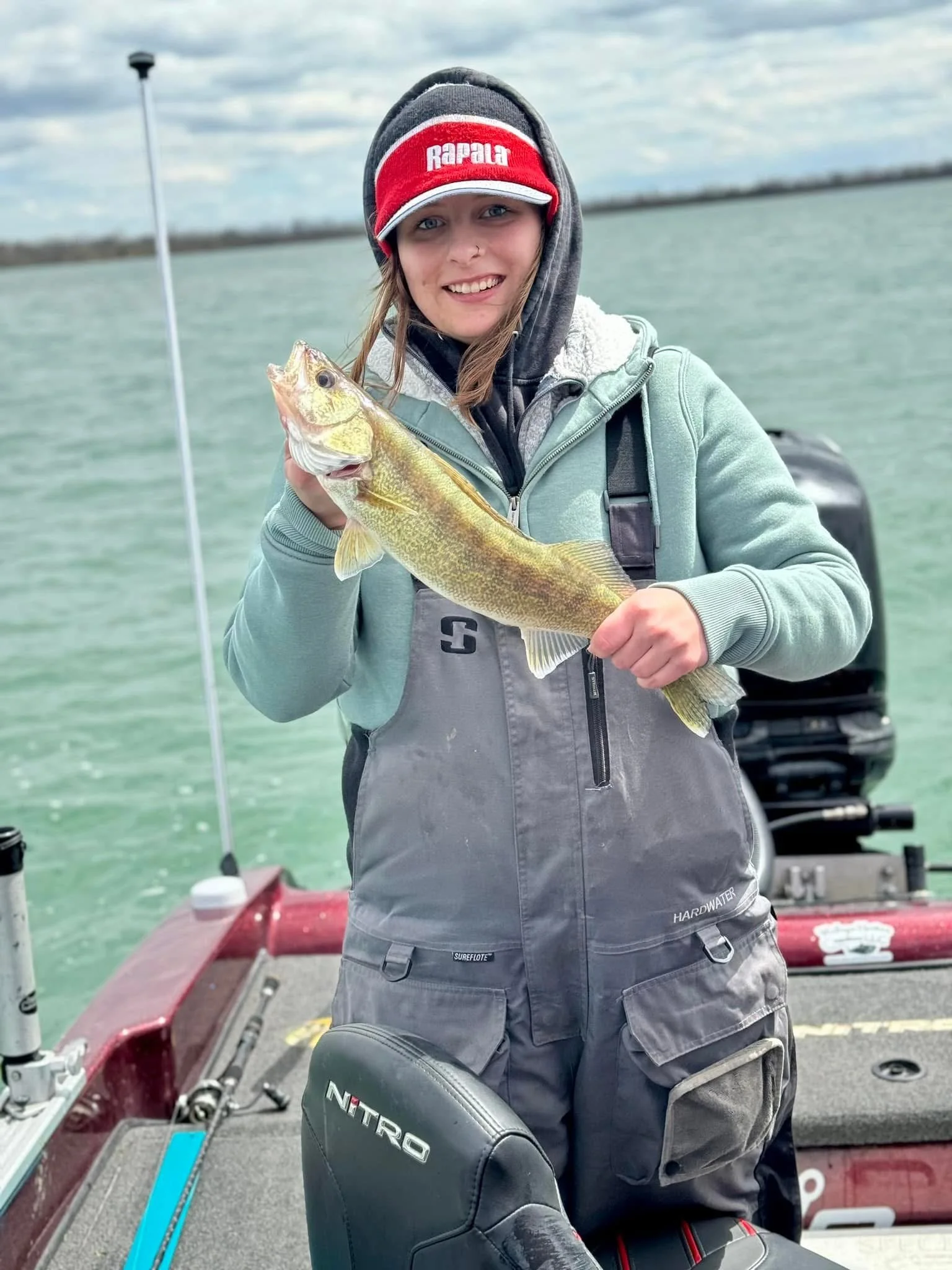 A young woman with a smile, wearing a red and gray 'Rapala' cap, gray hoodie, and gray overalls, holds a yellowish fish on a boat by the water with an outboard motor and cloudy sky in the background.