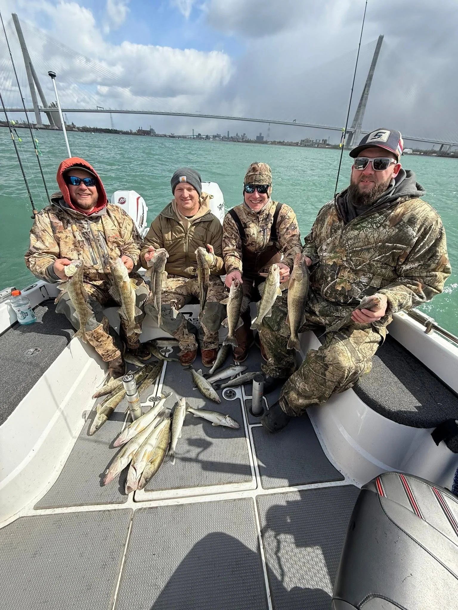 Four men sitting on a boat holding large fish with an architectural suspension bridge and city skyline in the background.
