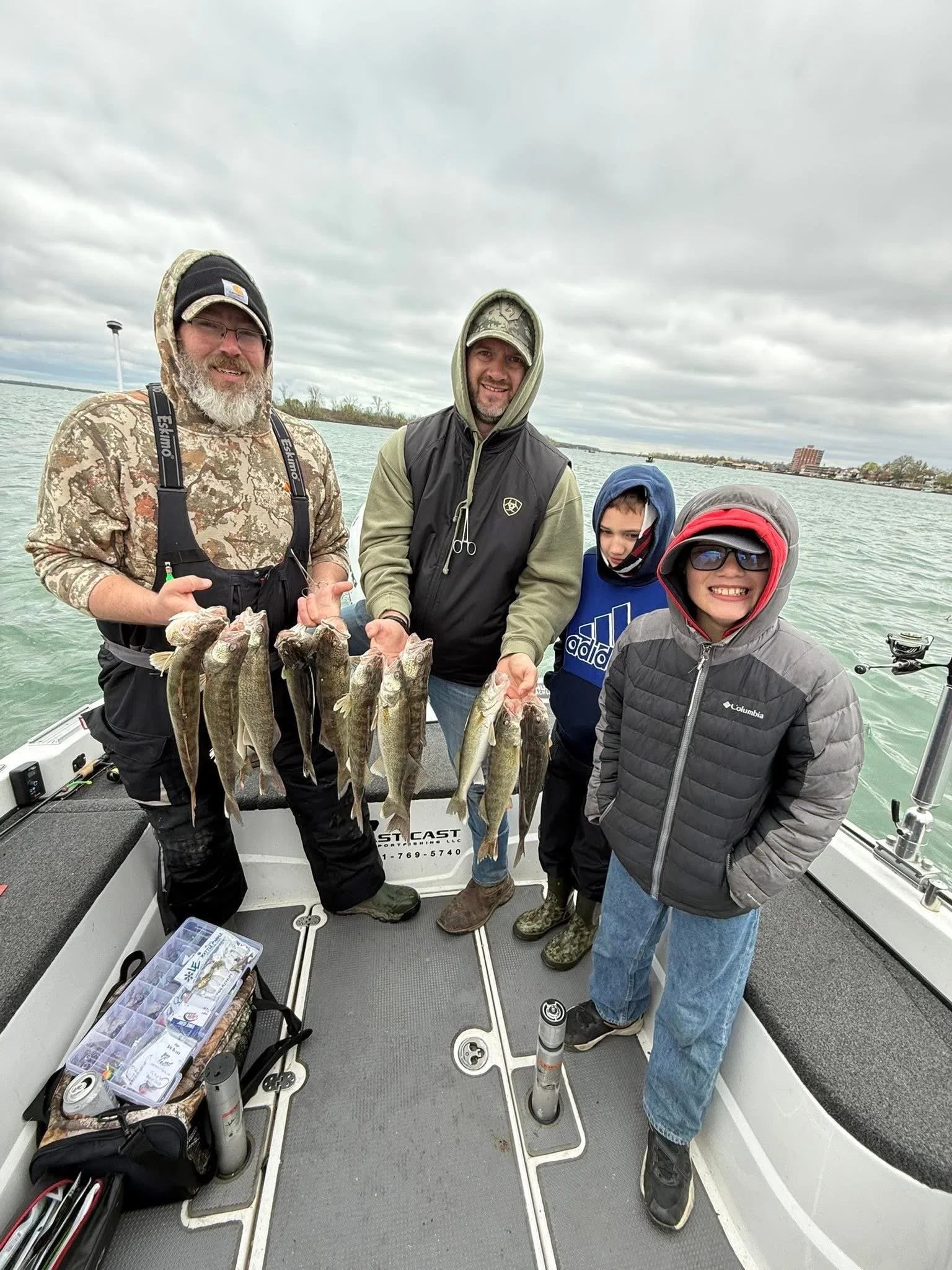 Four people on a boat with several fish hanging from a line, holding fishing gear and smiling at the camera, with cloudy skies and a body of water in the background.