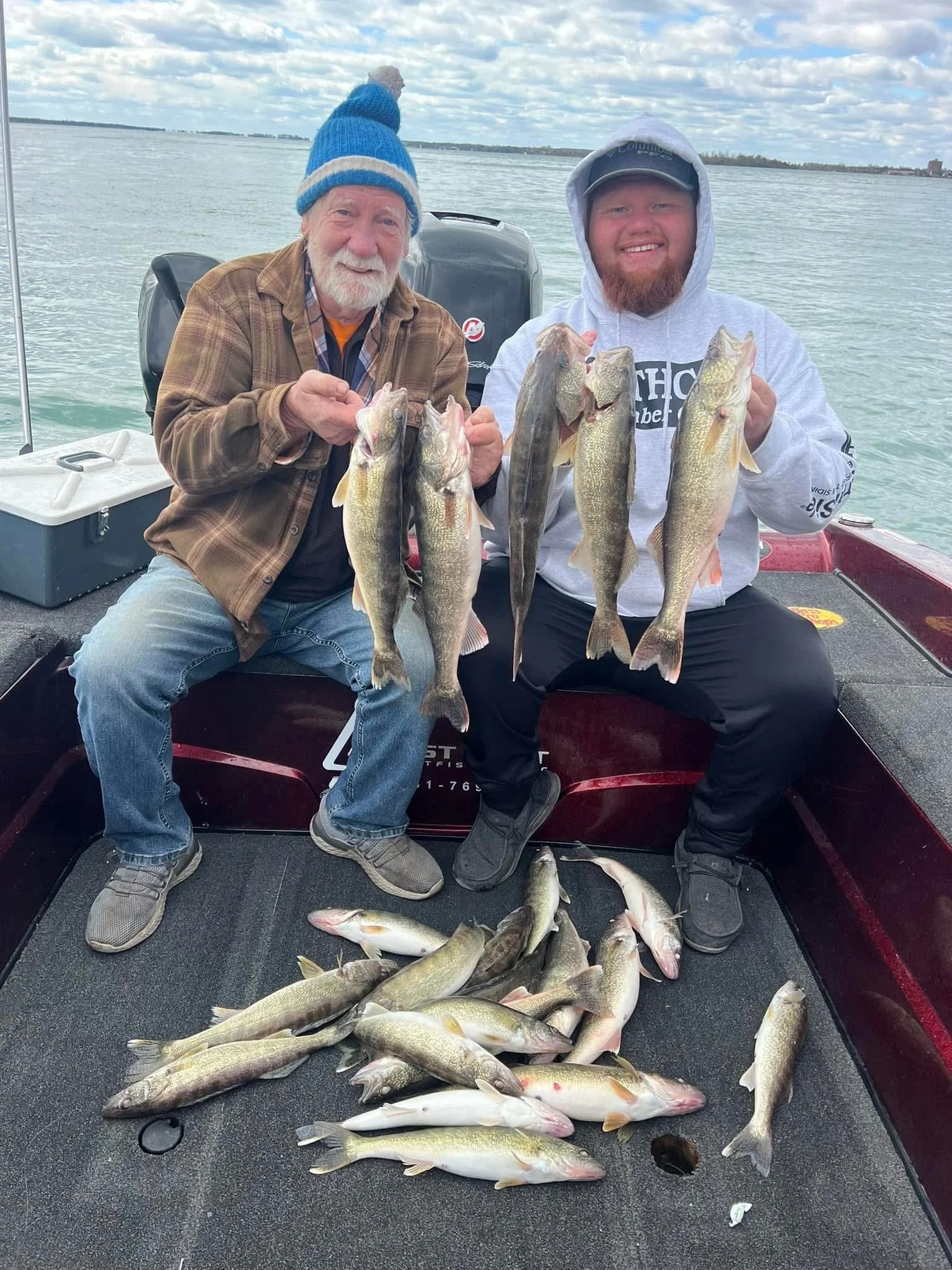 Two men on a boat holding multiple fish they caught, with more fish laid out on the boat floor, and a body of water and cloudy sky in the background.