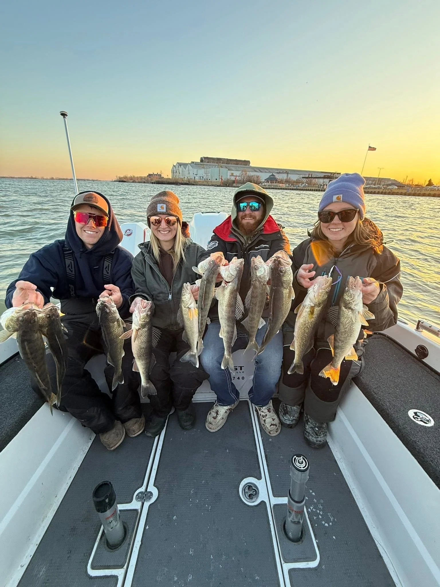Four people on a boat holding up caught fish at sunset with industrial buildings and water in the background.