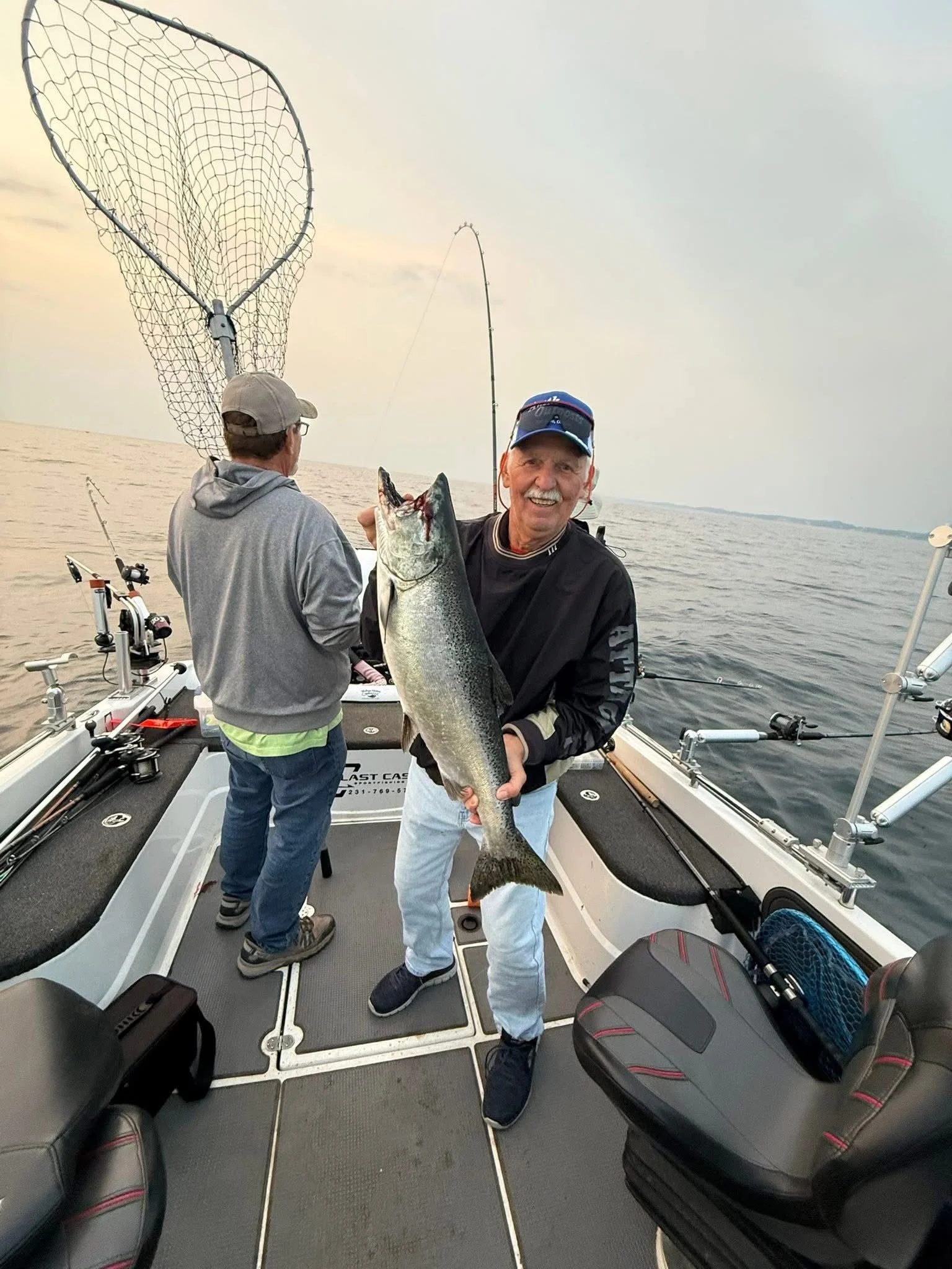 A man holding a large fish on a boat with another person in the background, fishing gear and equipment around, water and sky in the background.