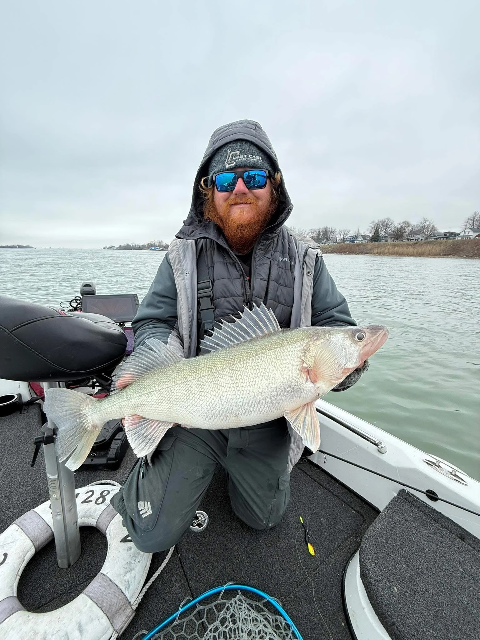 A man with a red beard, sunglasses, and winter clothing on a boat, holding a large fish he caught.