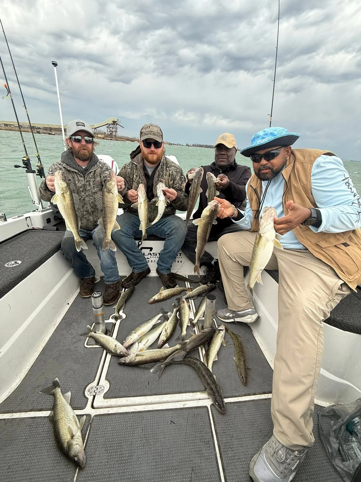 Four men sitting on a boat holding fish they caught, with more fish laying on the boat's floor. The sky is cloudy and there is water and industrial structures in the background.