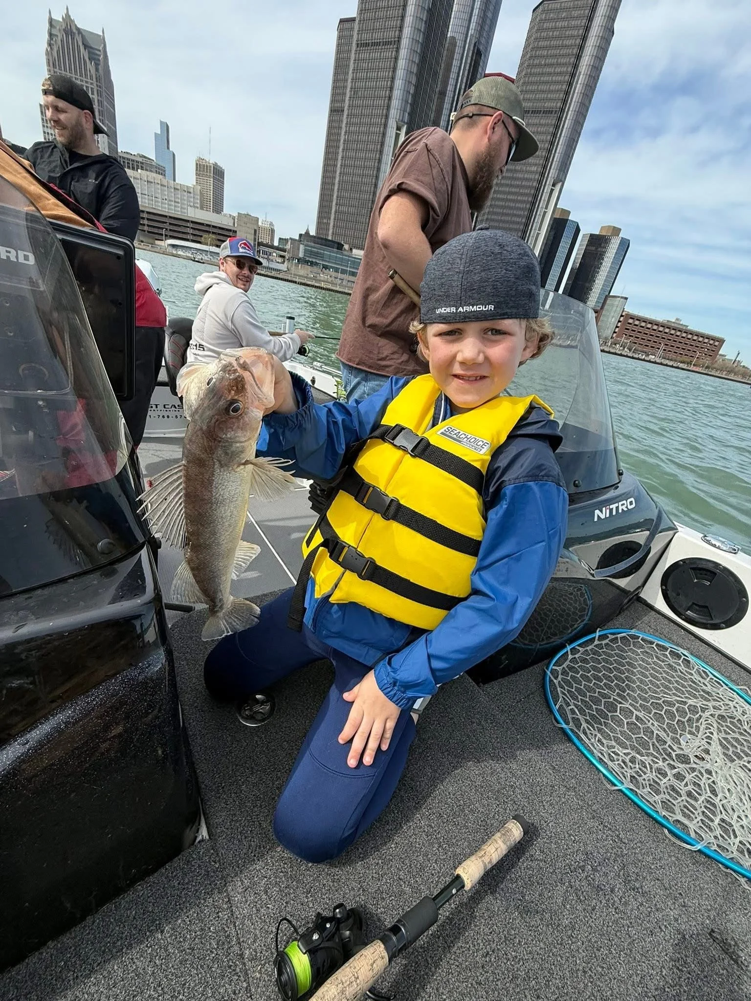 A young boy wearing a gray Under Armour cap, yellow life jacket, and blue jacket, holding a fish on a boat with city buildings in the background. Two men are nearby, one with a beard and glasses, and the other smiling and wearing sunglasses and a cap. Fishing gear is on the boat.
