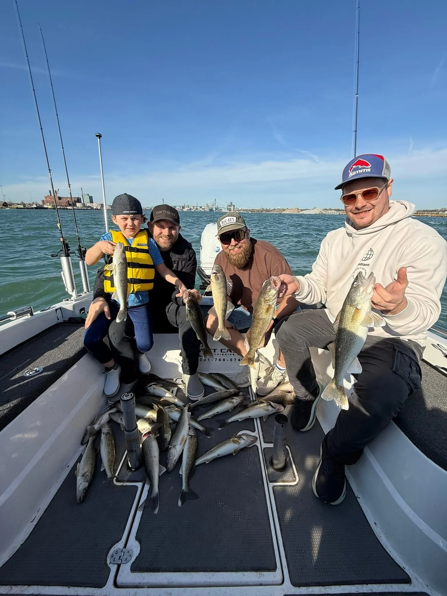 Four men with fishing gear on a boat holding multiple fish they caught, with a city skyline and bridge in the background, under clear blue skies.