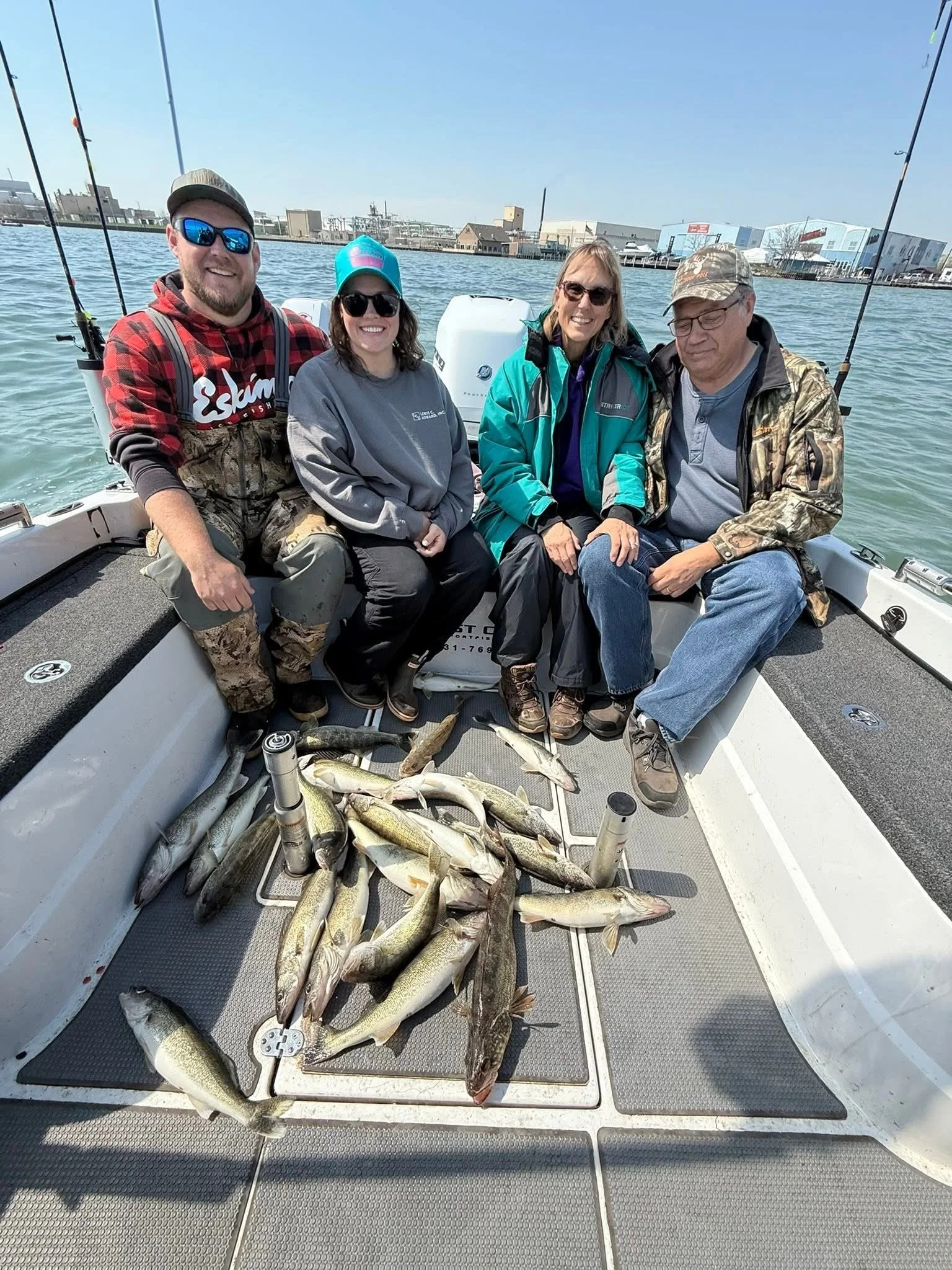 Four people sitting on a boat with a catch of multiple fish laid out in front of them, water and buildings in the background.