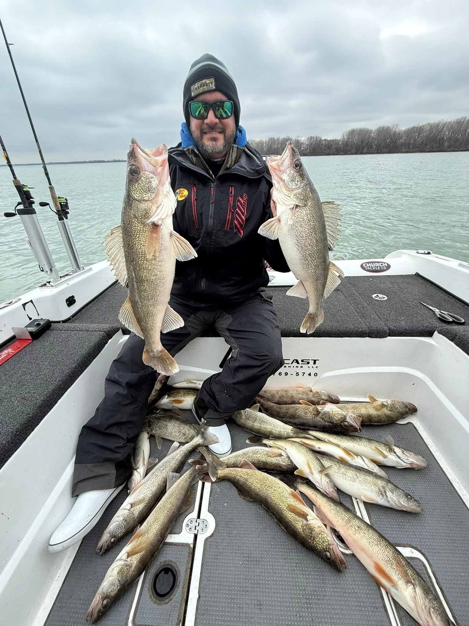 Man in a jacket and sunglasses on a boat holding two large fish, surrounded by many other fish on the boat floor, with water and cloudy sky in background.