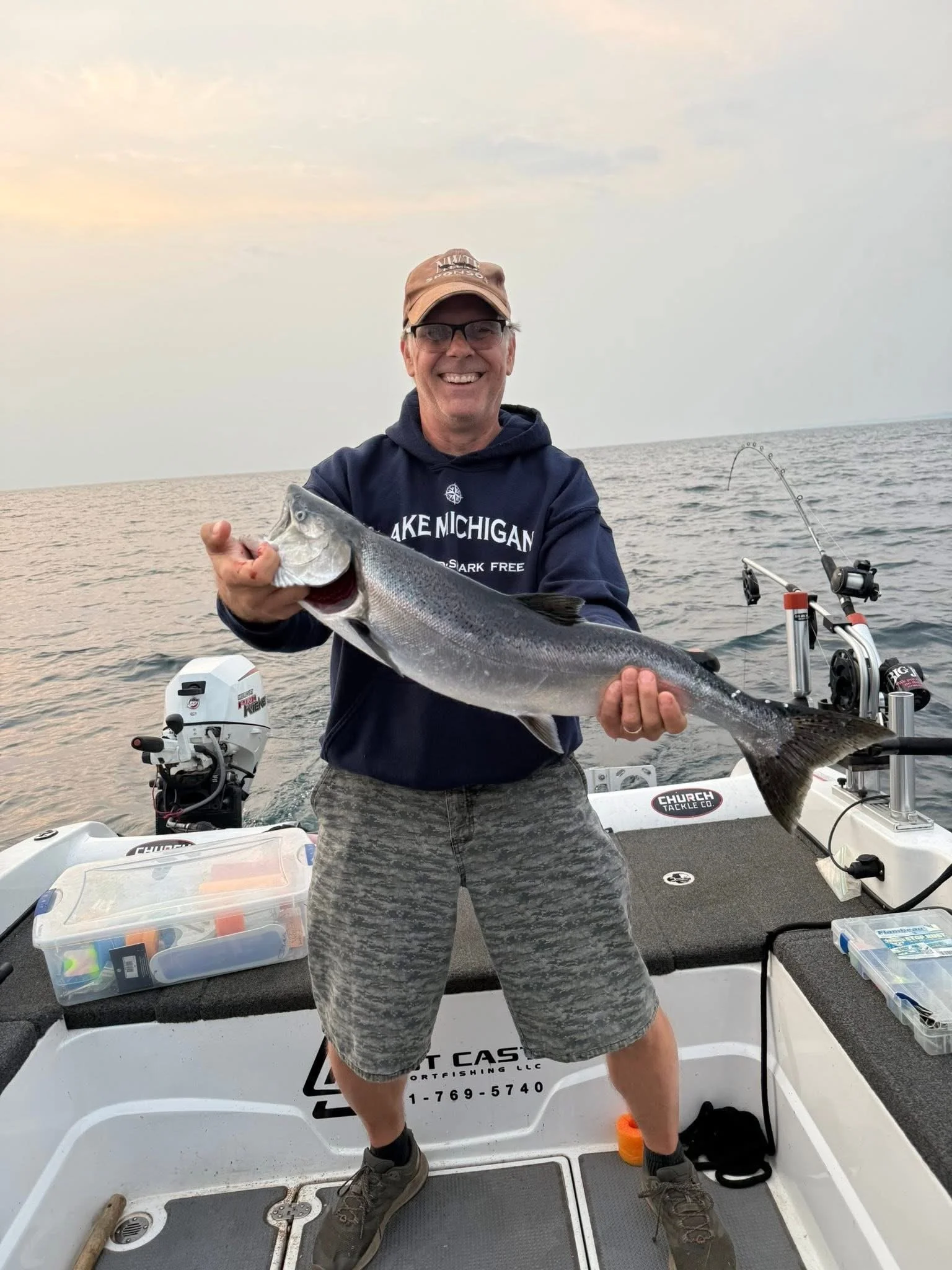 A man smiling and holding a large fish on a boat in the open water during sunset. The boat is equipped with fishing gear and storage boxes, and the man is wearing glasses, a cap, a hoodie, and camo shorts.