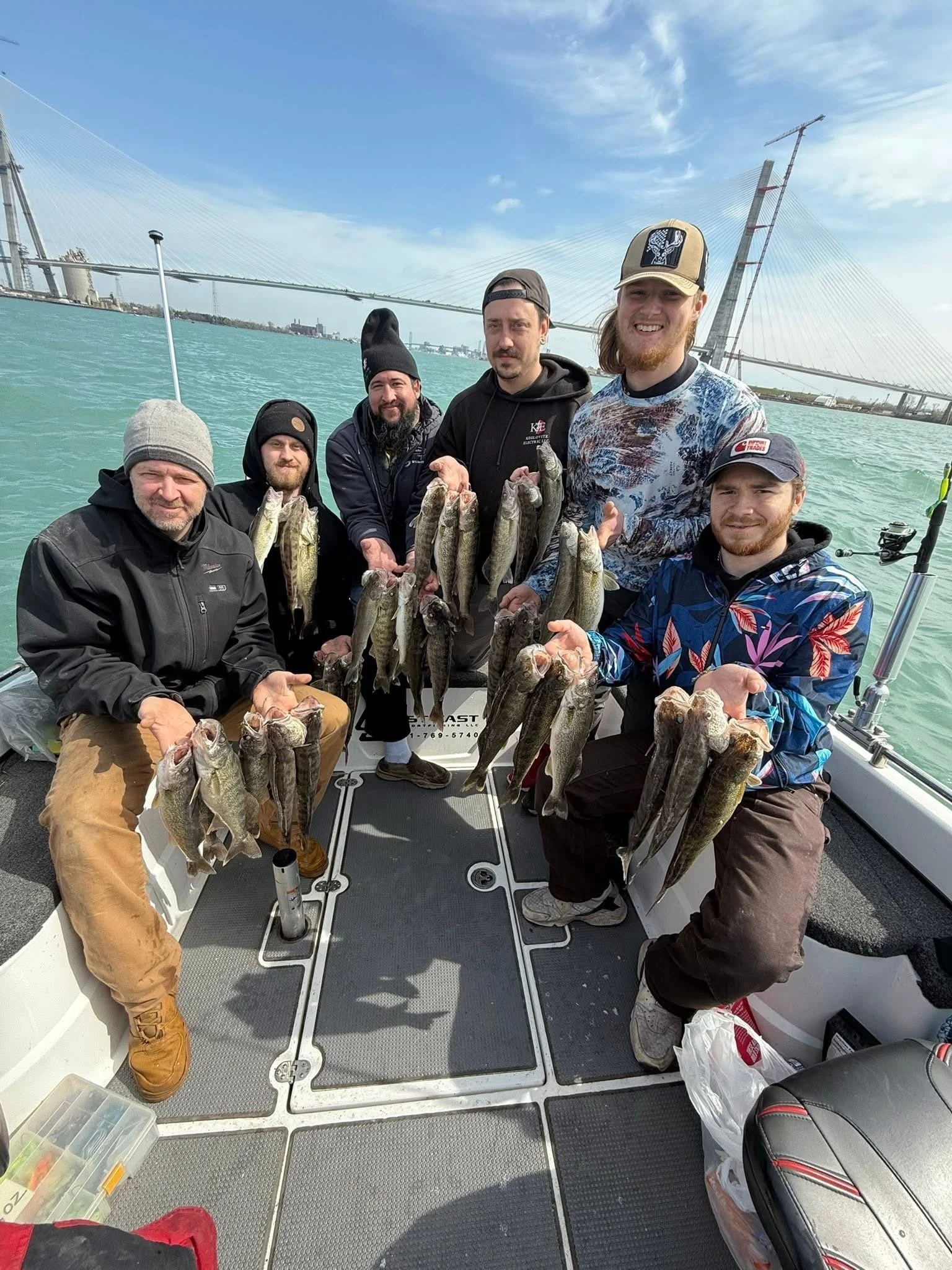 Group of six men on a boat holding fish they caught, with a bridge and blue sky in the background.
