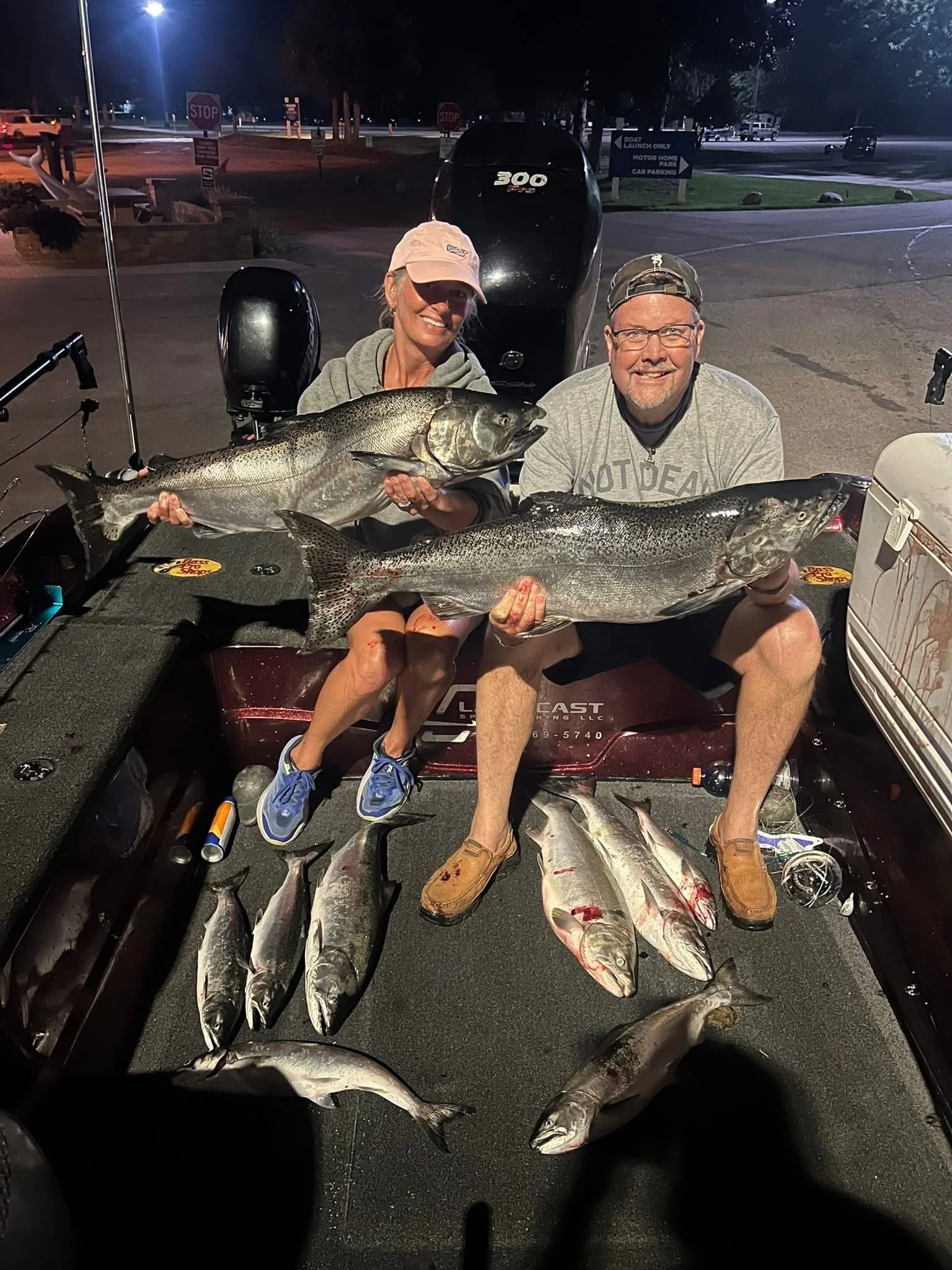 Two people sitting on a boat at night holding large fish with smiling faces, surrounded by additional fish on the boat's surface, with a dark outdoor background and vehicles in the distance.