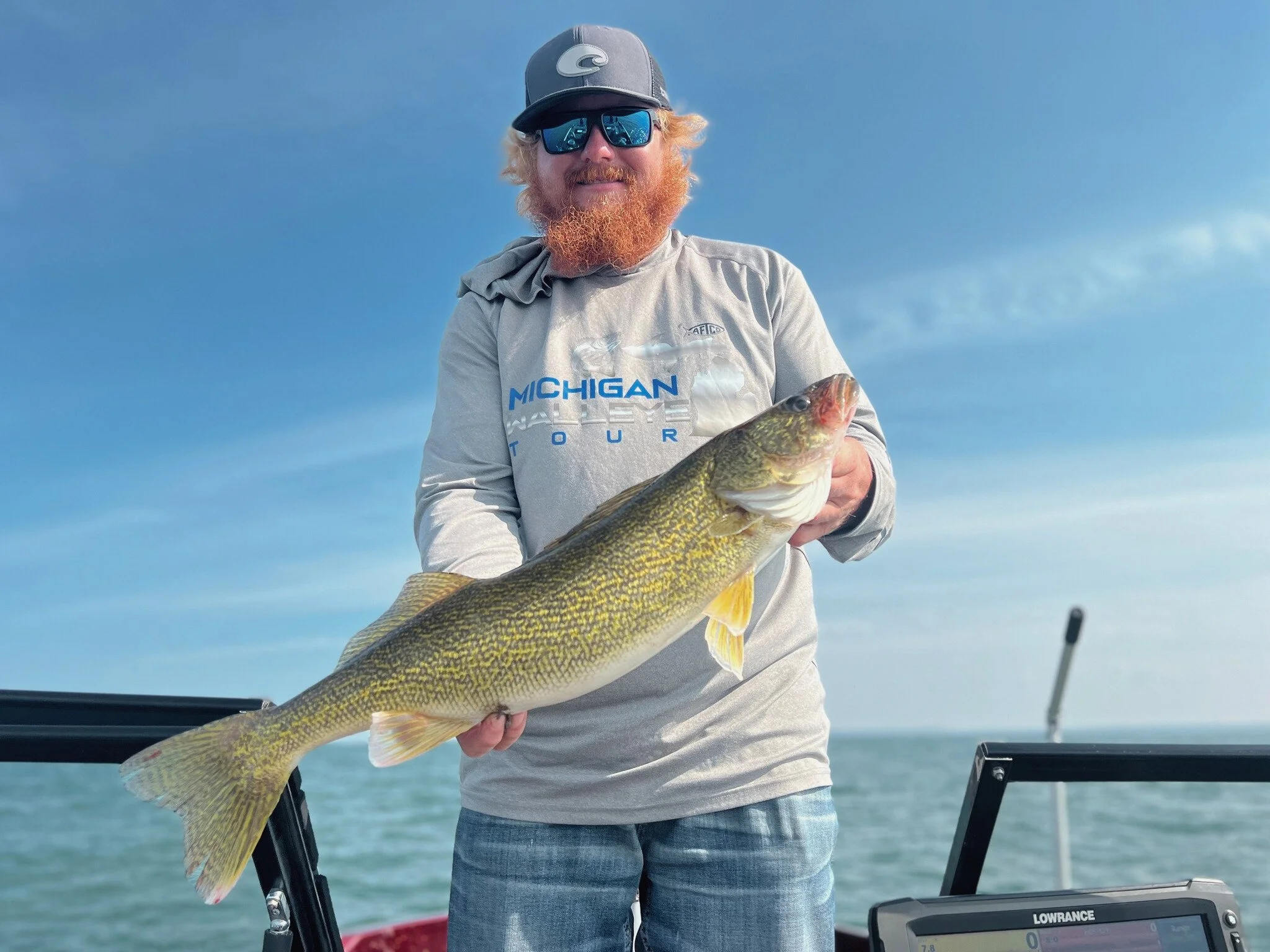 A man with a red beard wearing a cap and sunglasses holding a large fish on a boat during daytime. The background shows a clear sky and water.