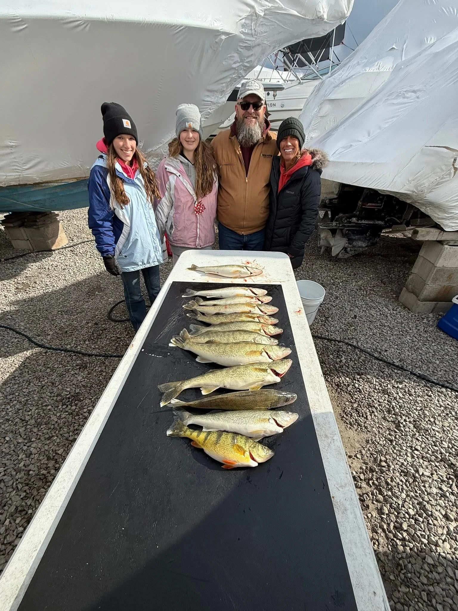 A group of four people standing behind a table with seven fish laid out on it. They are outdoors near boats covered with tarps. The group is smiling and dressed in warm clothing and hats.