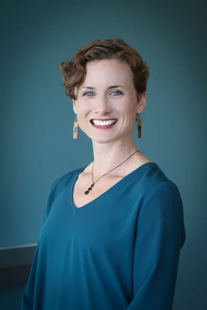 A smiling woman with short curly hair, wearing a teal blouse, dangling earrings, and a black necklace, standing against a blue-green background.