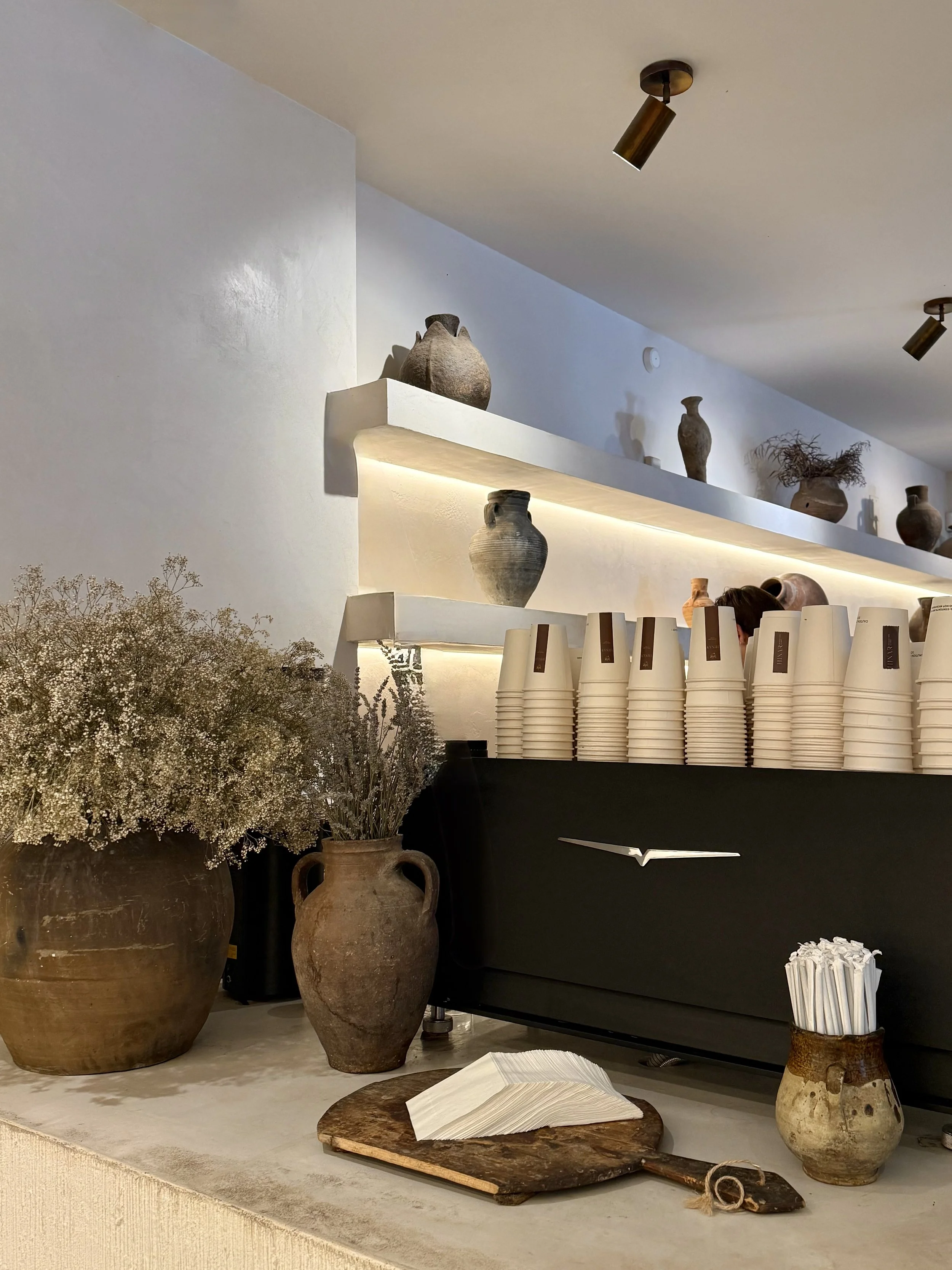 Decorative pottery vases and dried flowers on a counter with stacked disposable cups and a coffee machine in the background.