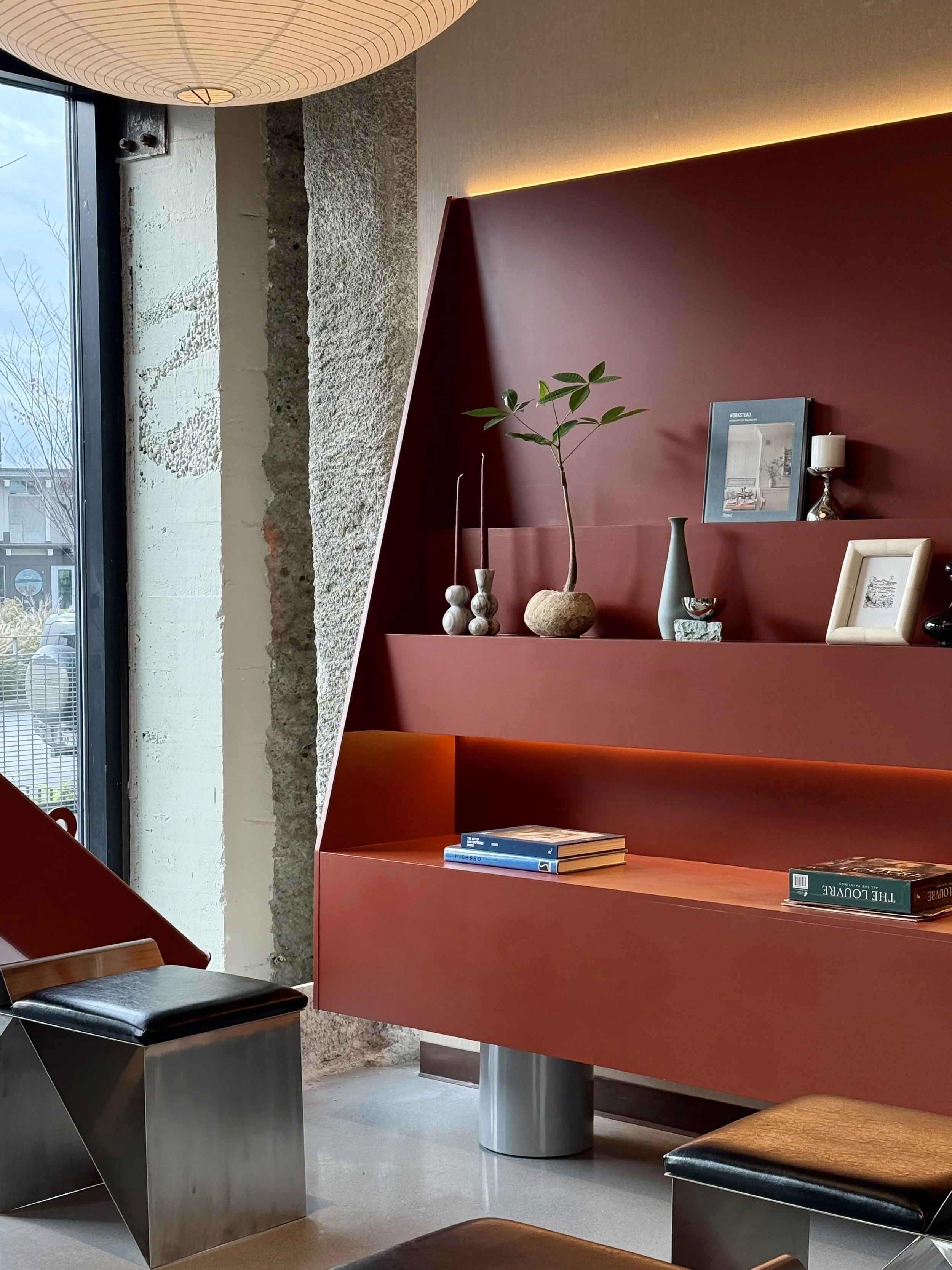 Interior of a modern room with a red wall unit displaying decorative items, vases, books, and a framed photo, next to a large window with a view outside.