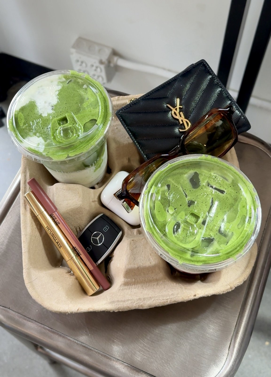 Two cups of green matcha drinks with ice, a black YSL wallet, sunglasses, a perfume lipstick, a Mercedes-Benz car key, earphones, and a tube of lip gloss on a beige cloth on a trolley.