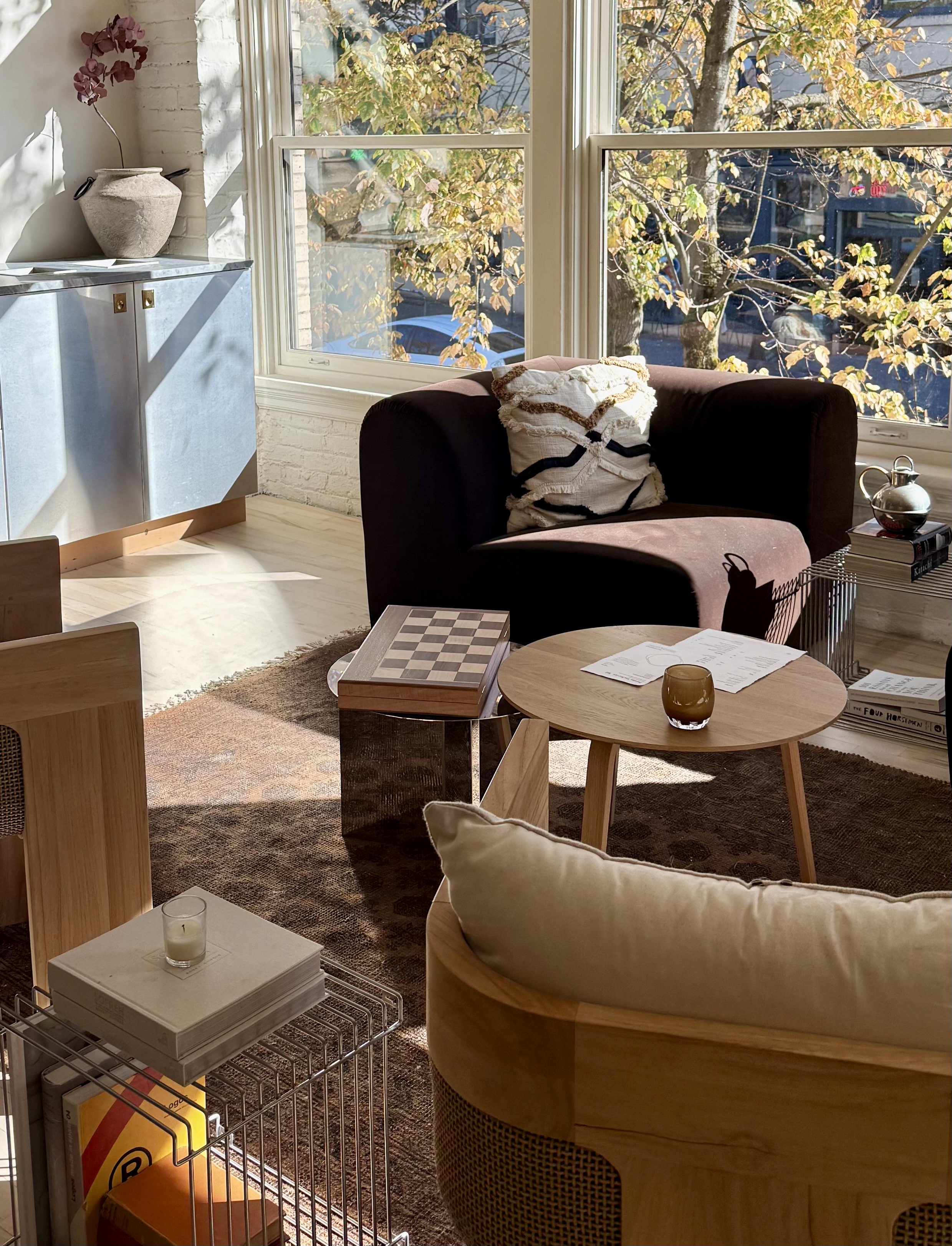 Sunlit living room with large windows, a dark brown sofa with a patterned pillow, a light wooden coffee table with papers and a glass, a round beige armchair with a white cushion, a side table with a teapot and books, a blue cabinet with a large vase and purple flowers, and a metal wire basket with books and a candle on top.