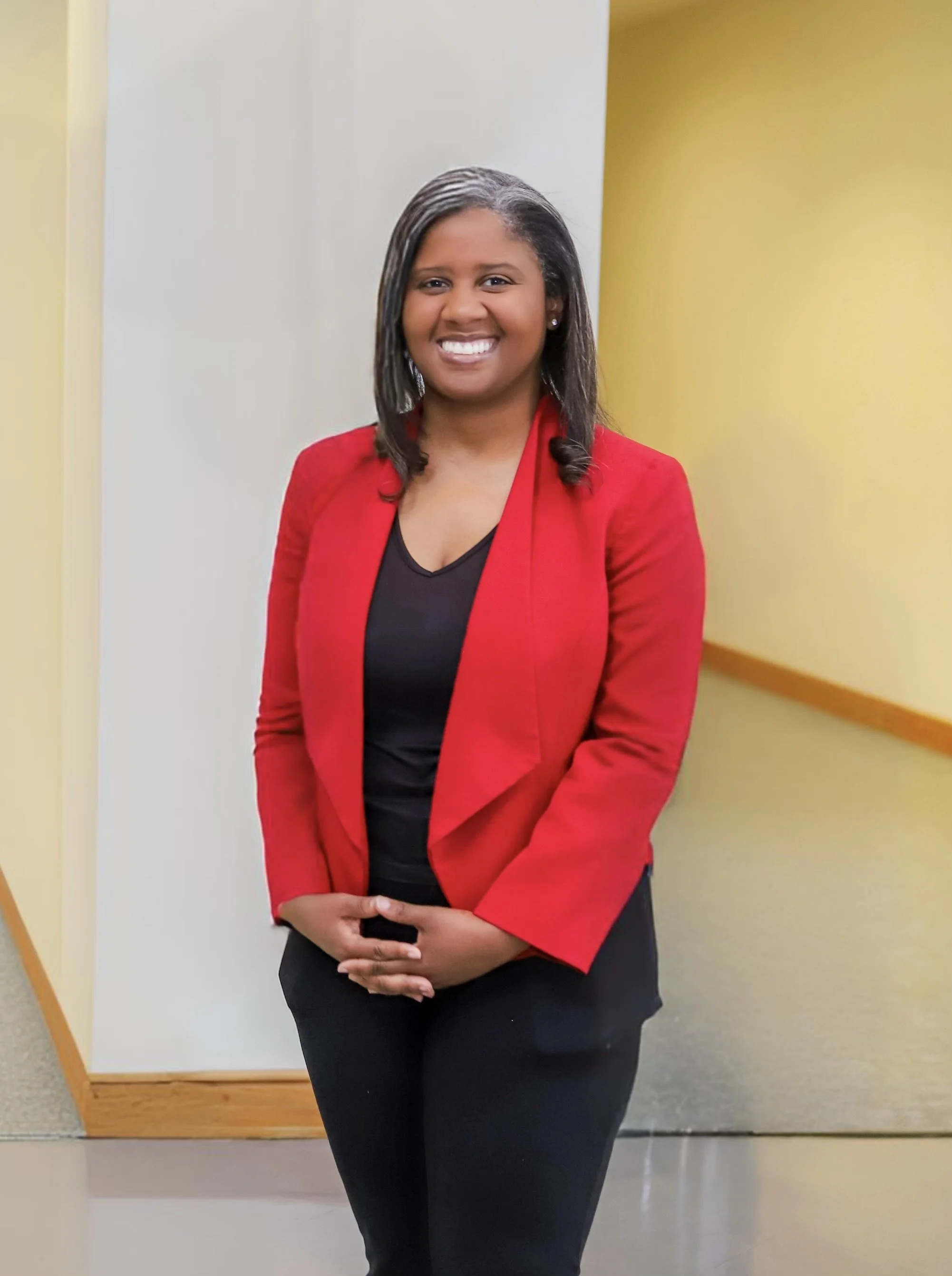 Margaret Franklin wearing a red blazer and black top, smiling and standing indoors against a background of white, yellow, and wooden wall panels.