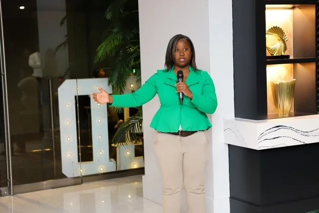 A woman in a green jacket holding a microphone and speaking in an indoor setting.