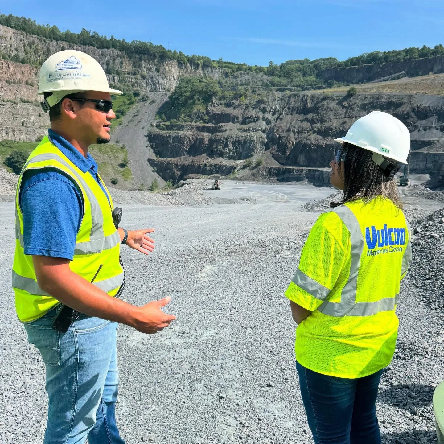 Margaret Franklin with a construction worker in safety vests and hard hats having a conversation at a gravel quarry with steep rock walls and a small heavy machinery in the background.