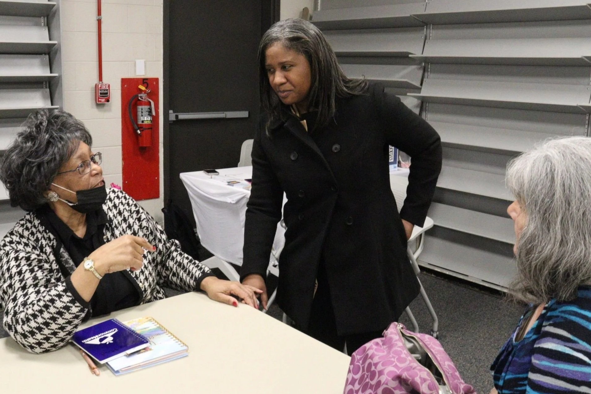 Three women engaged in conversation at a table in a conference room. One woman is standing, wearing a black coat, while the other two women are seated. The woman on the left is wearing glasses and a black and white patterned top with a face mask. The woman on the right has gray hair and is wearing a striped top. There are notebooks and a pink tote bag on the table, and a fire alarm and fire extinguisher are visible on the wall in the background.