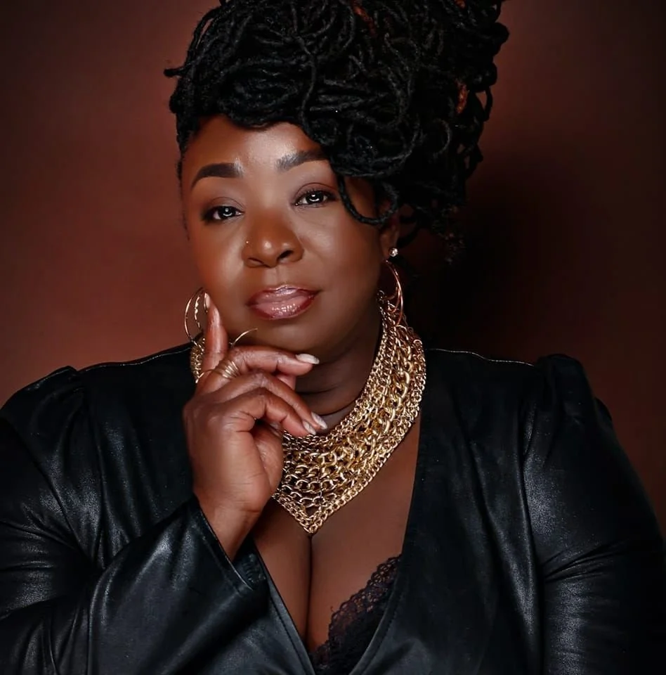 A woman with dark skin and voluminous dreadlocked hair styled to one side, wearing a black leather jacket and a layered gold chain necklace. She has hoop earrings, a nose piercing, and shiny lip gloss, striking a confident pose with her hand resting on her chin against a dark brown background.
