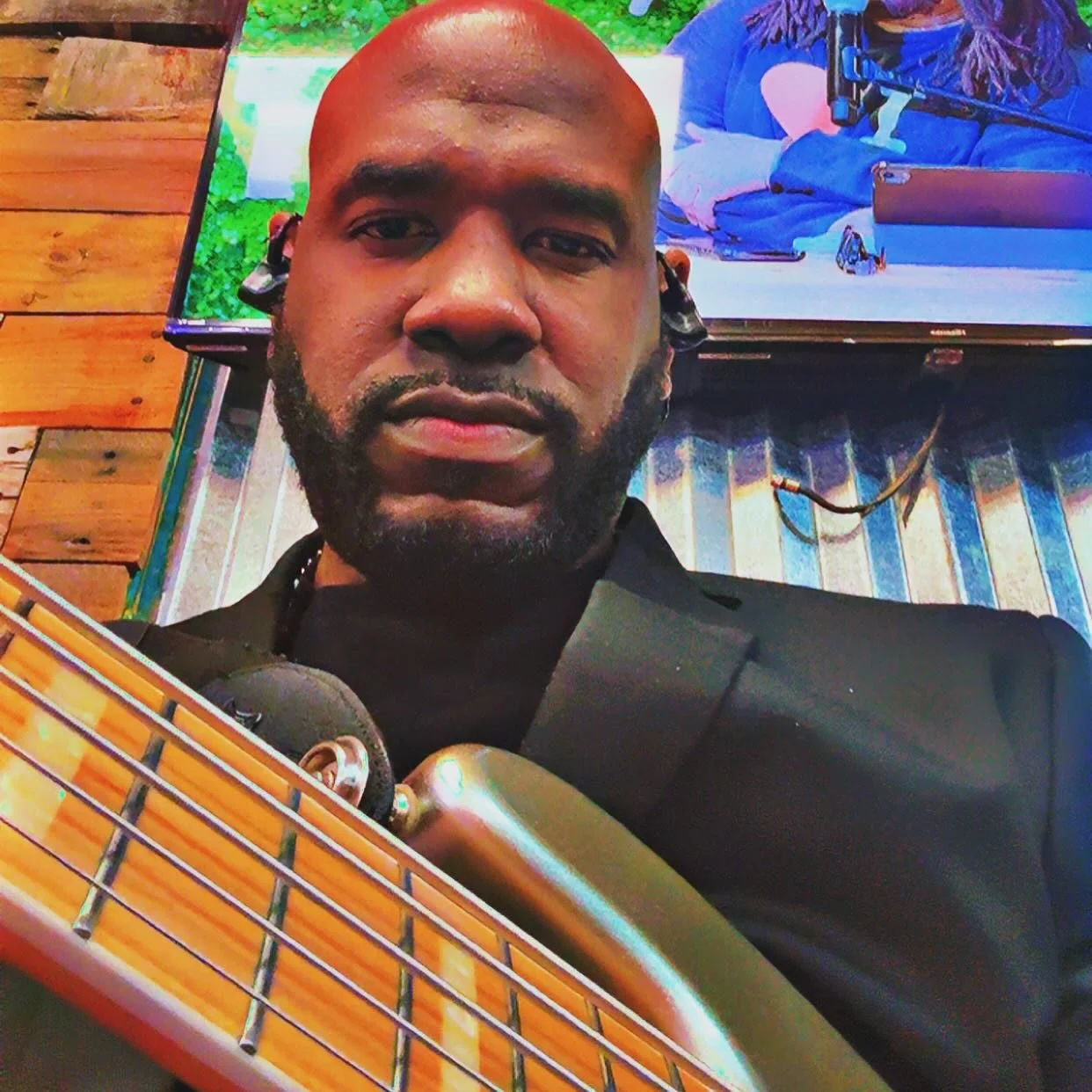 A man with a beard and bald head holding an acoustic guitar, wearing a black jacket and headphones; behind him is a colorful background with a large television screen showing a person with a gun and a laptop.