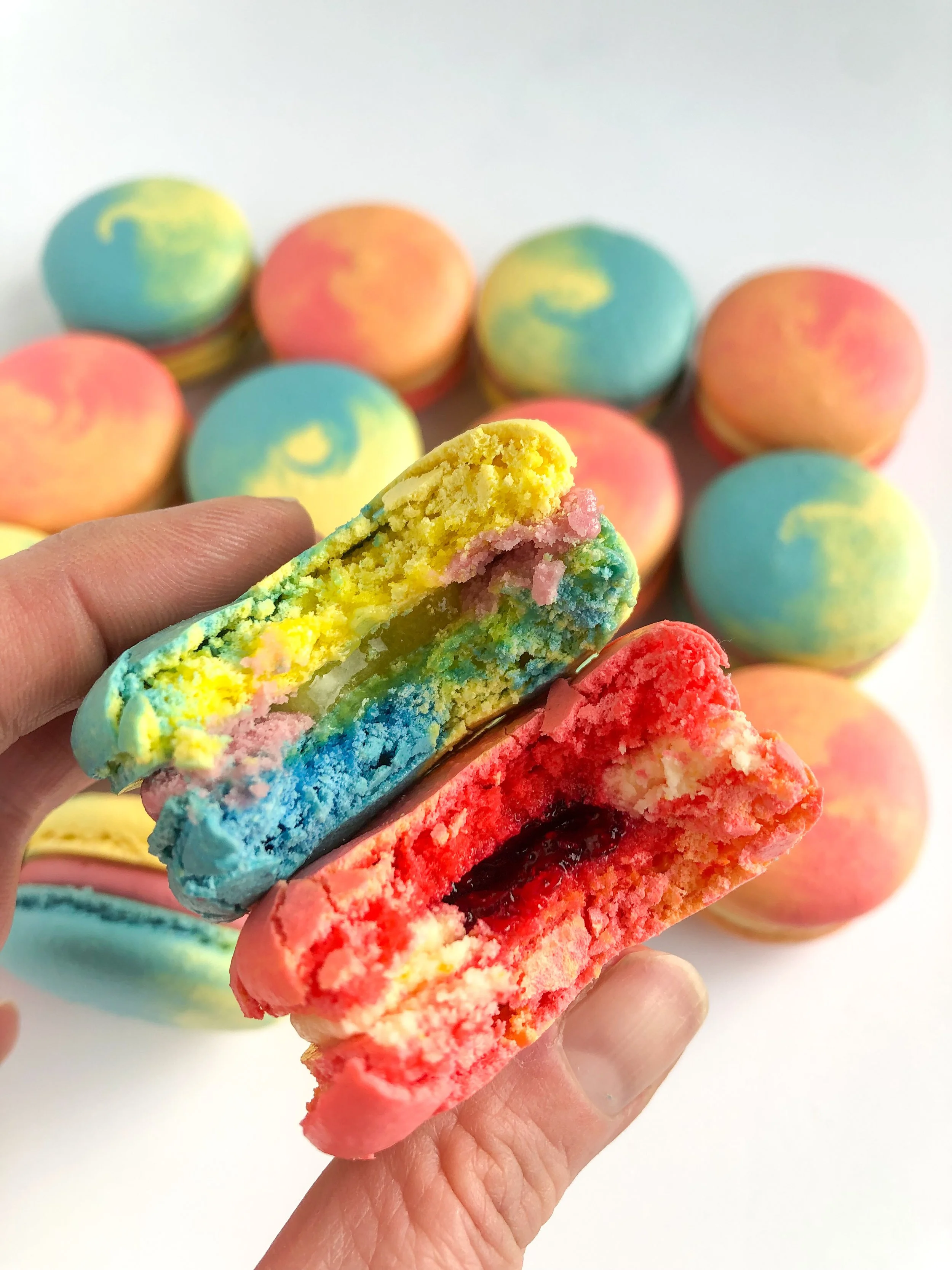 Close-up of a partially eaten rainbow-colored macaron with a fruity filling, held in hand, with several other brightly colored macarons in the background.