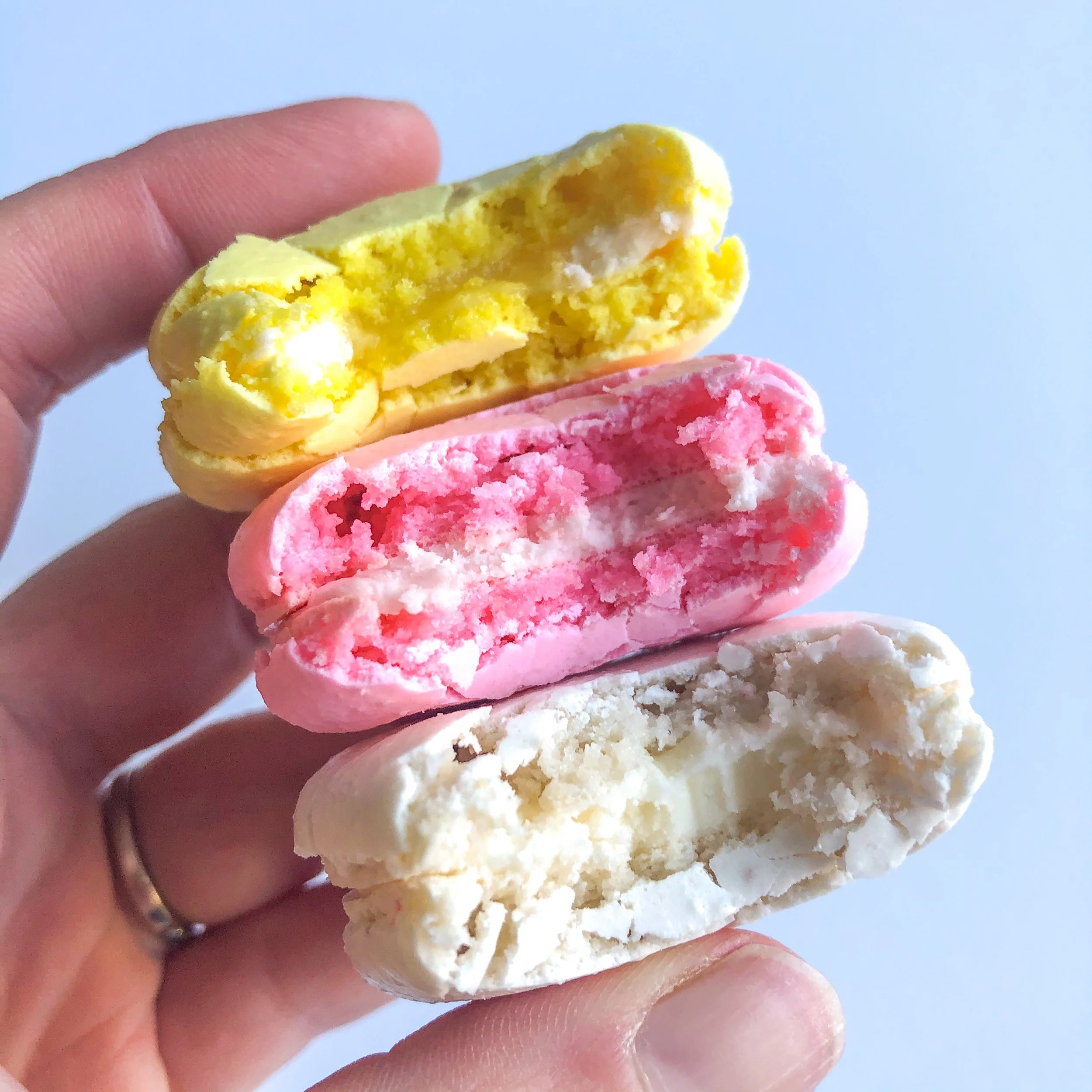 Close-up of three colorful macarons stacked vertically, held in a person's hand, against a plain light blue background.