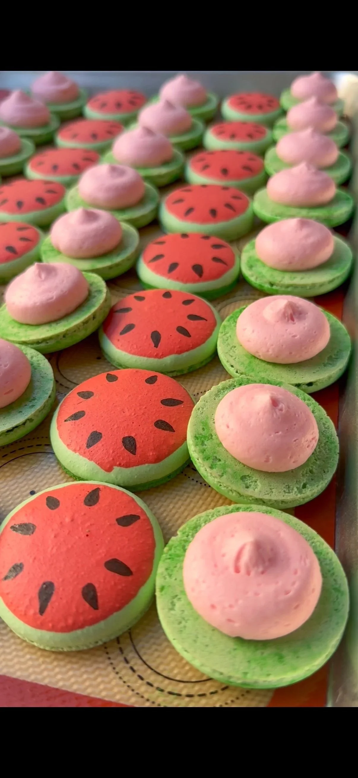 Macarons decorated to look like watermelons with pink frosting on top of green and red bases, arranged on a baking sheet.