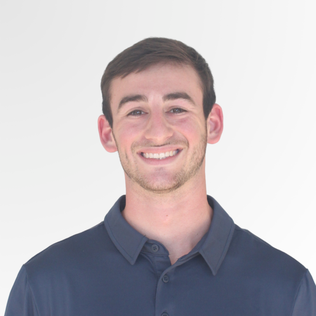 A smiling young man with short brown hair, wearing a navy blue collared shirt, standing against a plain white background.