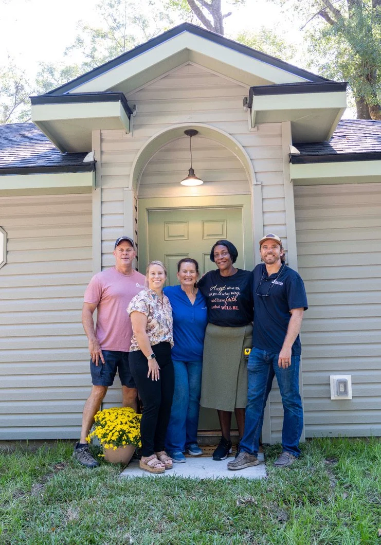 3 woman in the middle and 1 man on either side of the group smiling in front of a vinyl house with a cream white front door